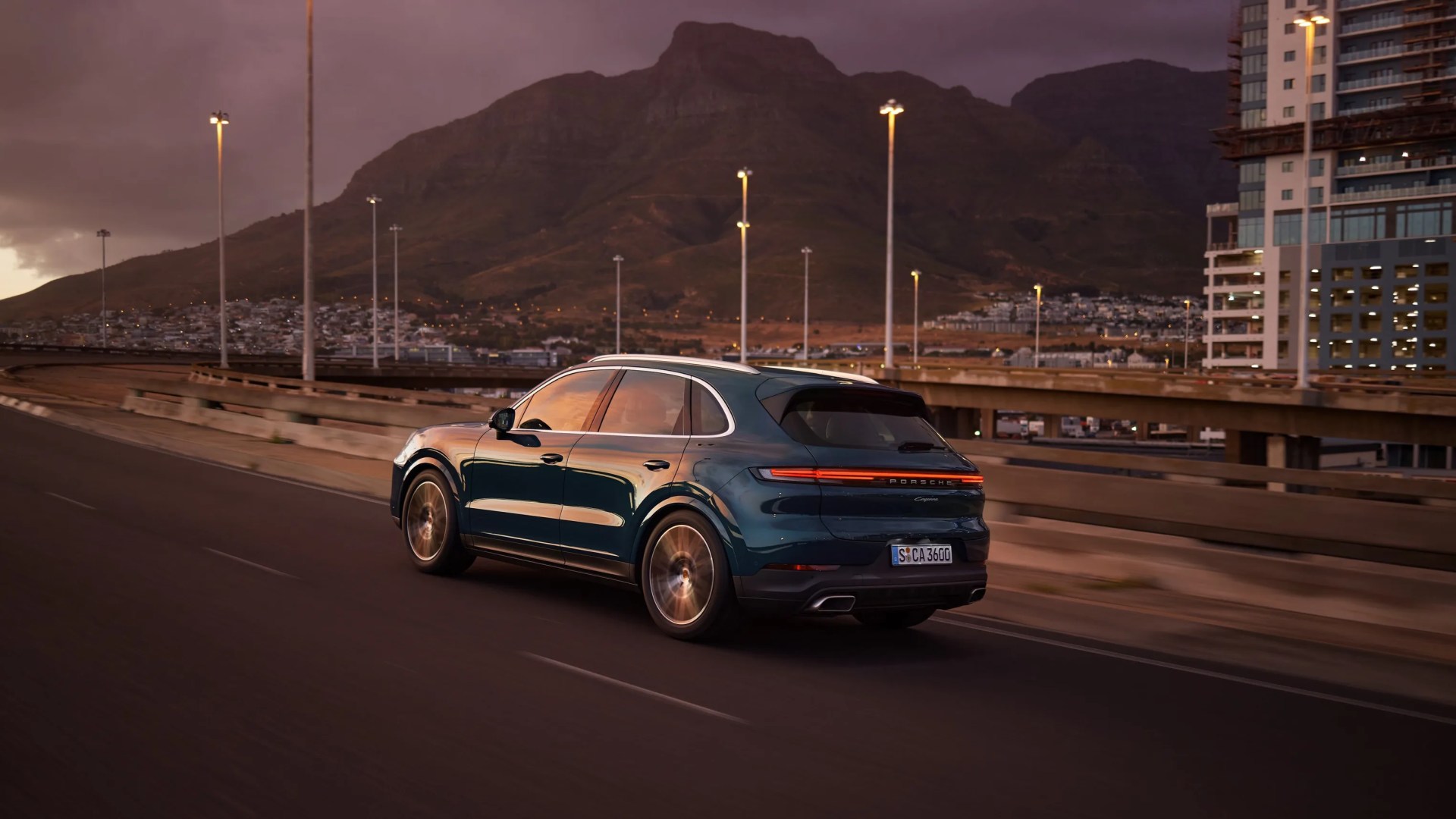 Dark green Porsche Cayenne SUV driving on a highway with mountains and city buildings in the background at dusk.