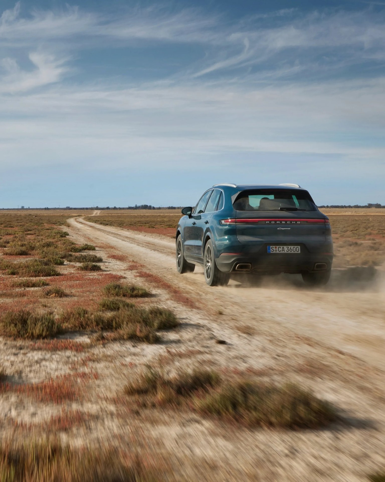 Blue Porsche Cayenne SUV driving on a dusty dirt road in a flat, open landscape under a partly cloudy sky.