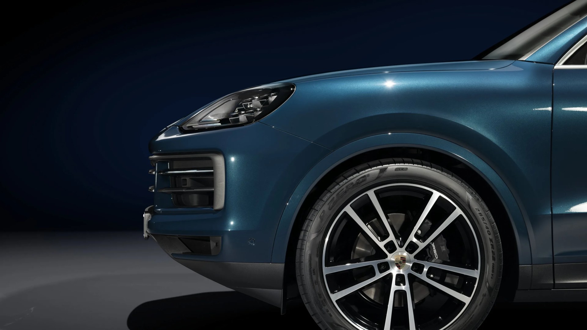 Front side view of a metallic blue Porsche SUV showing the headlight and black alloy wheel with silver spokes.