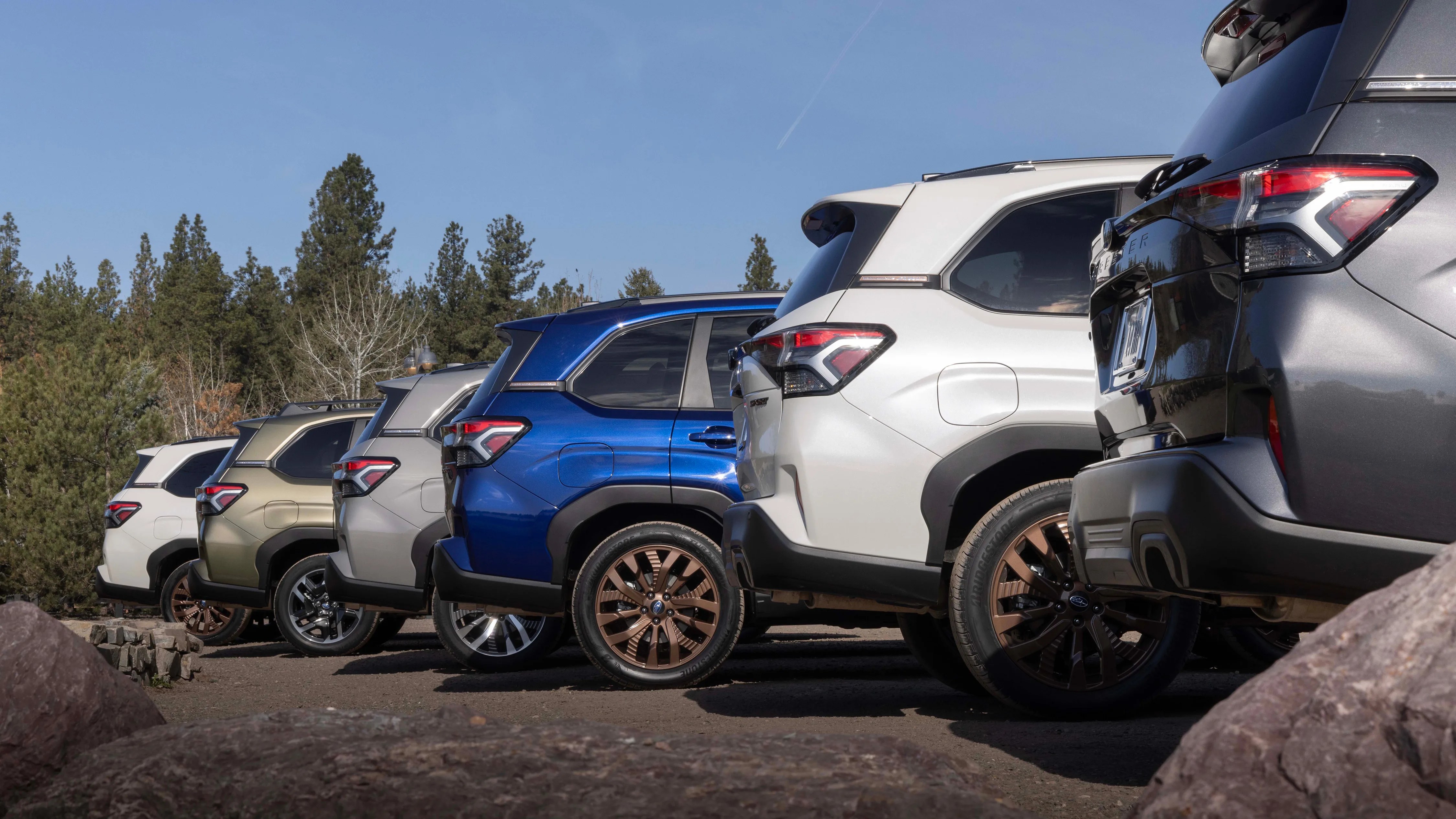 Five SUVs in white, bronze, silver, blue, and gray colors parked side by side on a dirt surface with trees in the background.