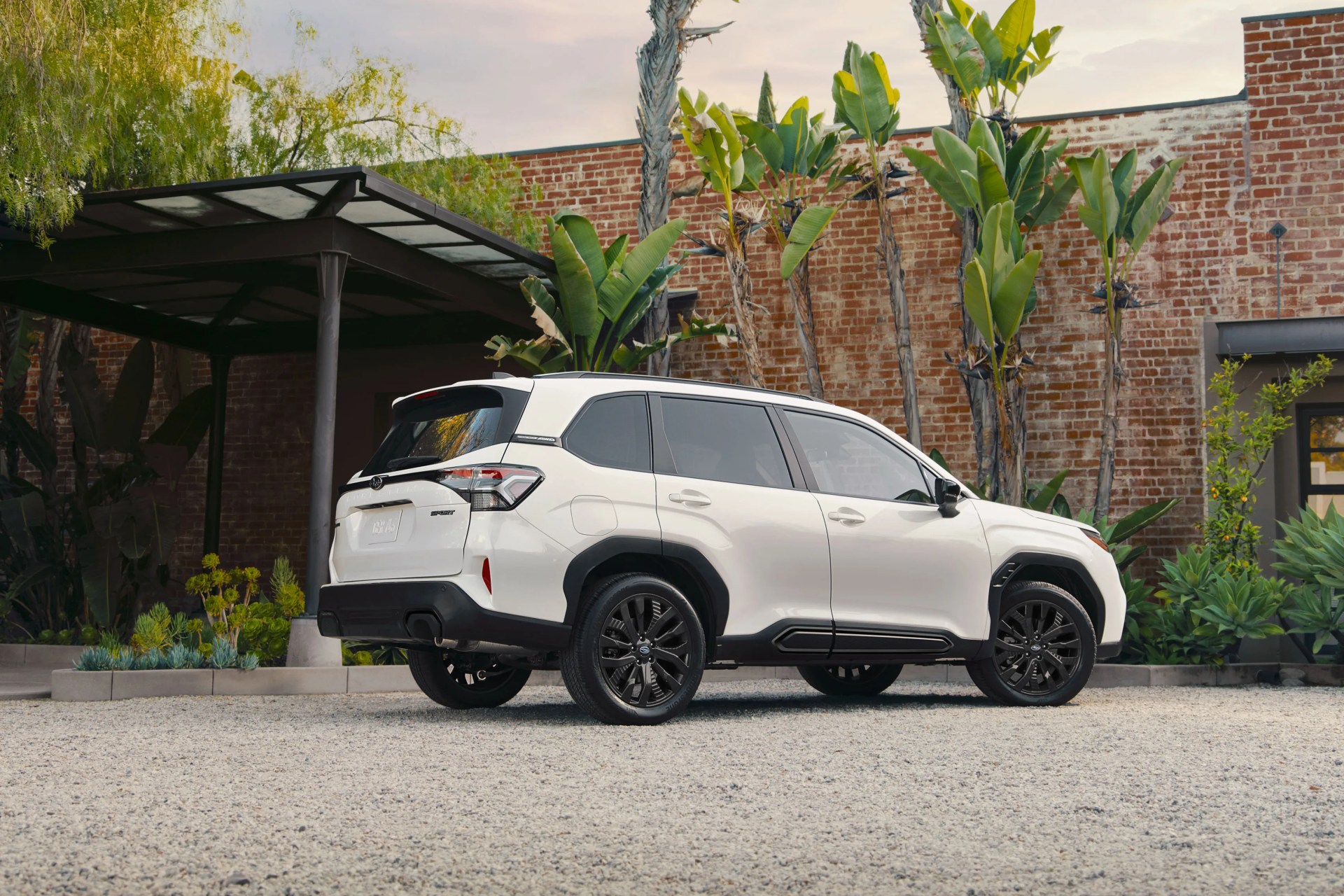 White SUV with black trim and black wheels parked on gravel in front of a brick wall with tropical plants.