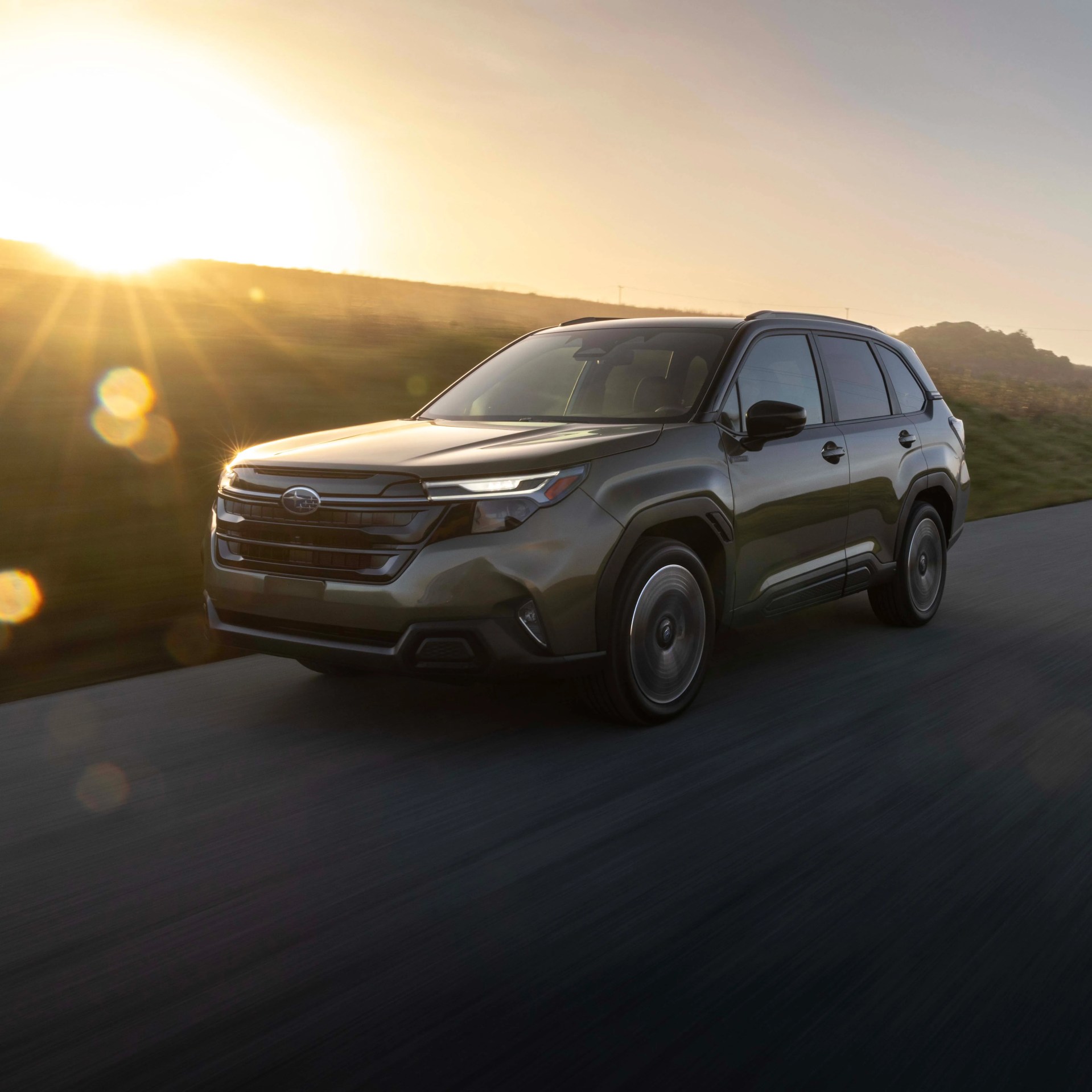 Dark green Subaru SUV driving on a road at sunset with sun flare in the background.