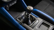 Black leather manual gear shift knob with silver top and blue stitching on black leather boot in a car interior.