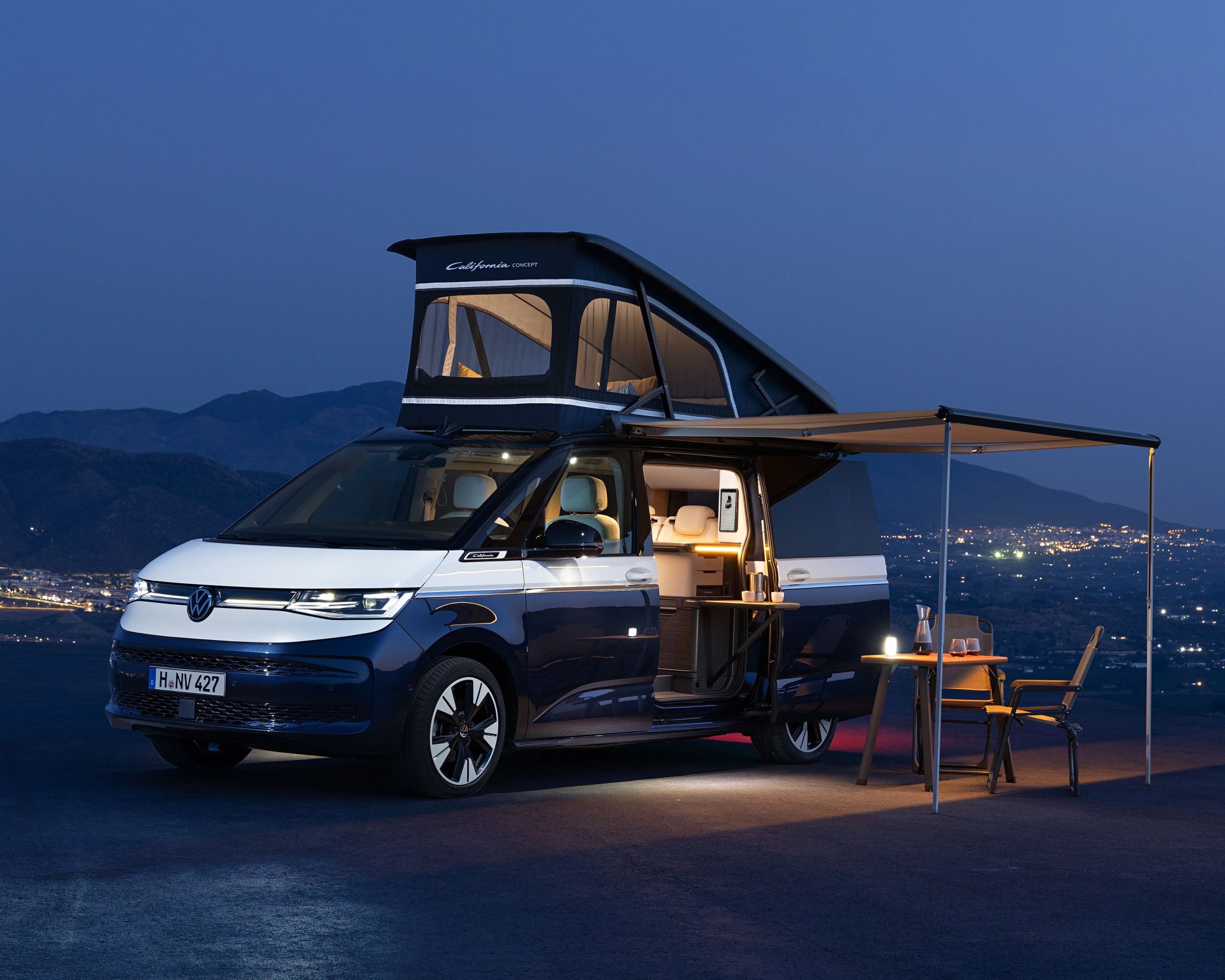 Blue and white Volkswagen camper van with pop-up roof and awning, parked at dusk with outdoor table and chairs.