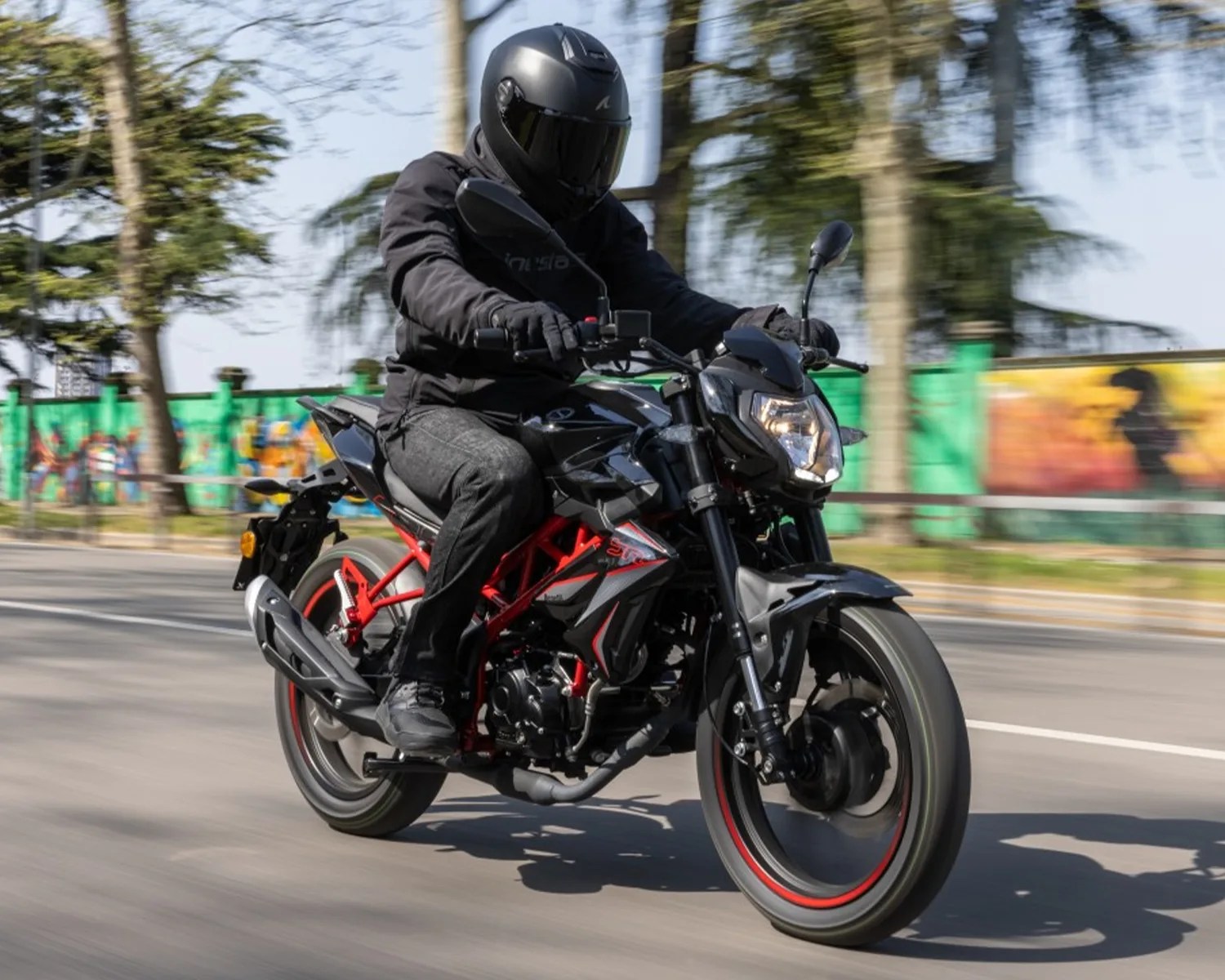 Person wearing black helmet and jacket riding a black and red motorcycle on a road with trees and graffiti in the background.