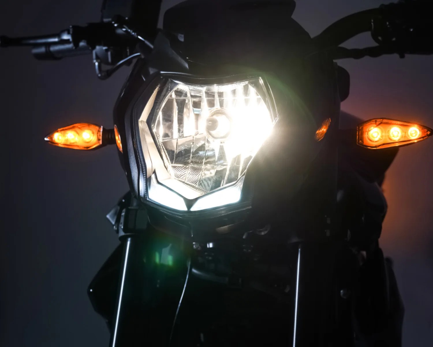 Front view of a motorcycle headlight and orange turn signals illuminated in a dark setting.