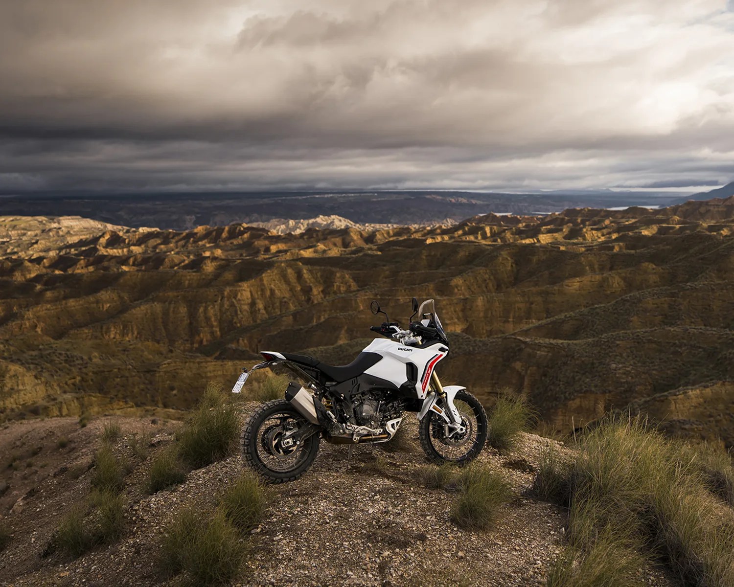 White and black Ducati adventure motorcycle parked on a rocky hill with rugged mountainous terrain and cloudy sky in the background.