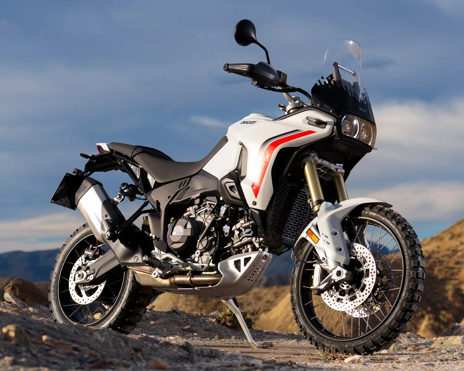 White Ducati off-road motorcycle with knobby tires parked on rocky terrain under a blue sky.