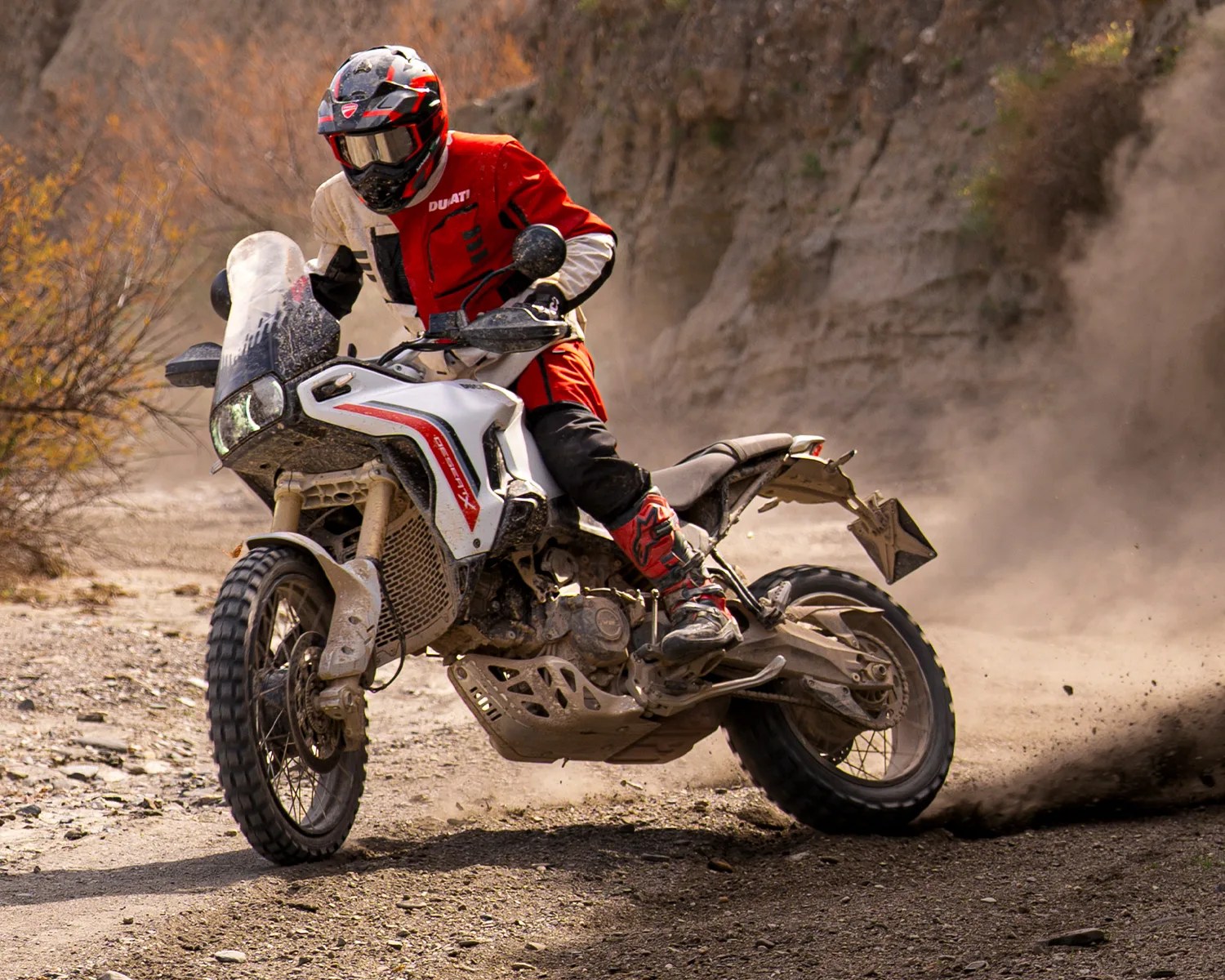 Dirt-covered white and red adventure motorcycle with rider in red and white gear kicking up dust on rocky trail.