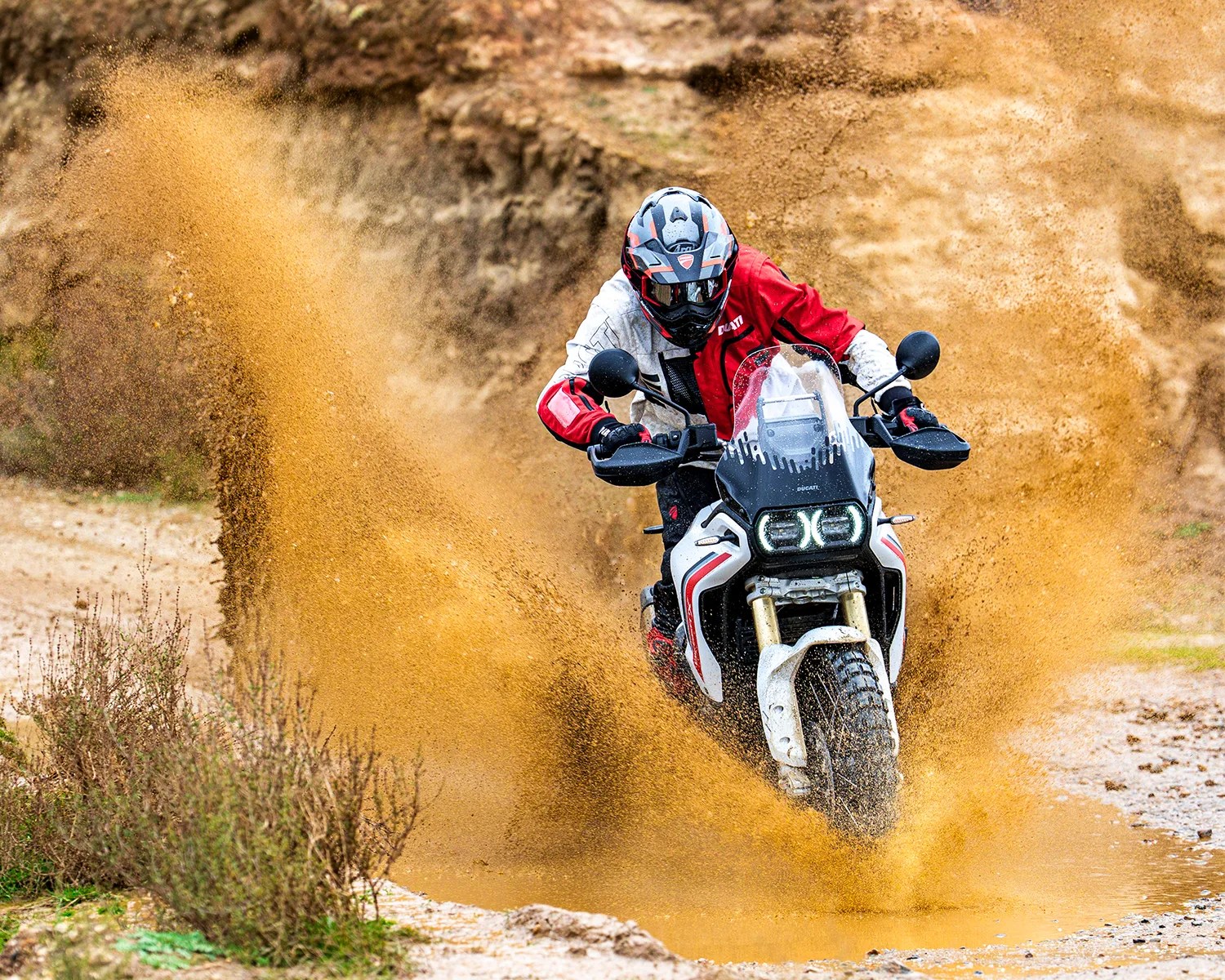 Motorcyclist in red and white gear riding a white and black Ducati off-road bike splashing muddy water.