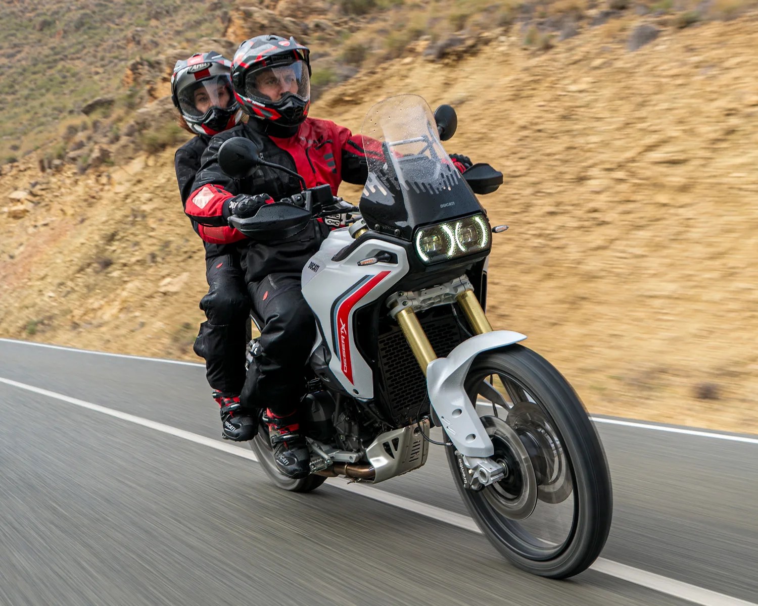 Two people wearing helmets and riding gear on a white Ducati DesertX motorcycle on a paved road.