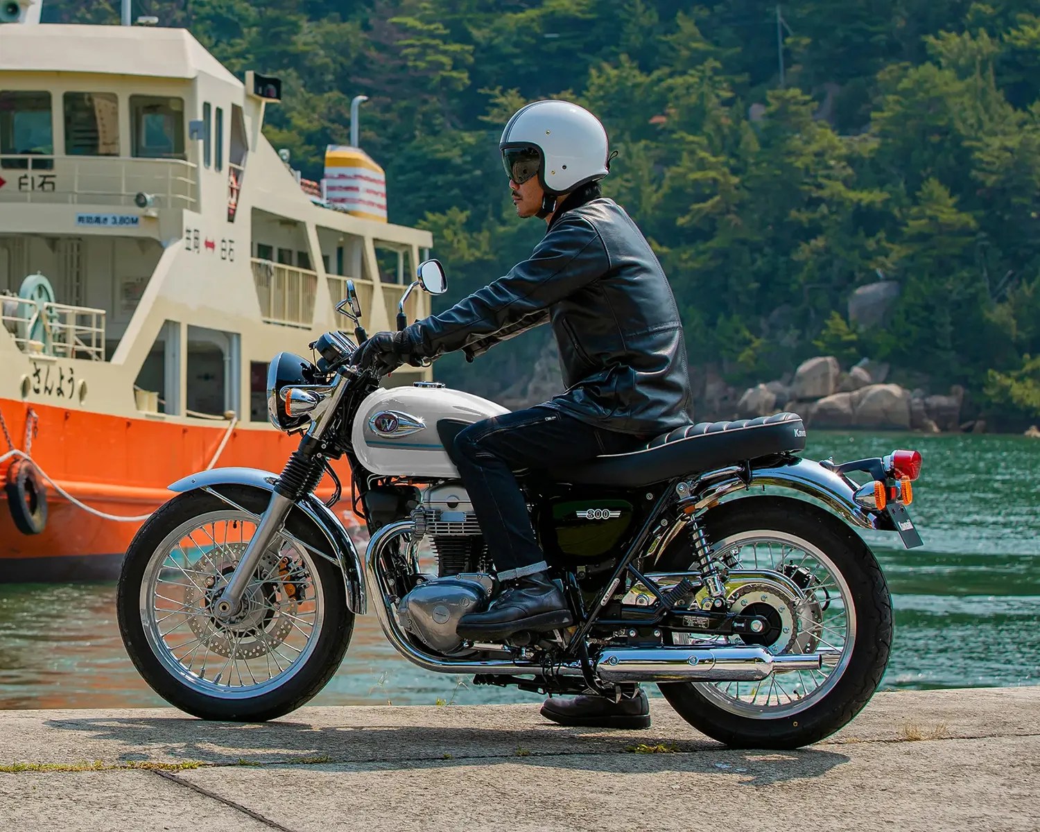 Person wearing a white helmet and black leather jacket sitting on a silver and black classic motorcycle by the water with a ferry boat in the background.