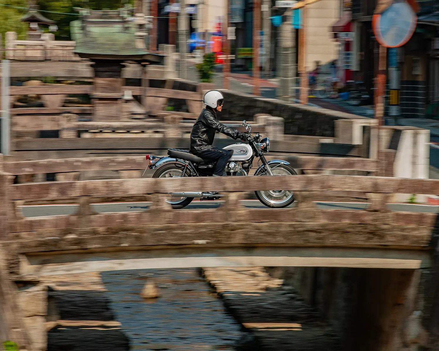 Person wearing a white helmet and black leather jacket riding a silver motorcycle across a stone bridge.