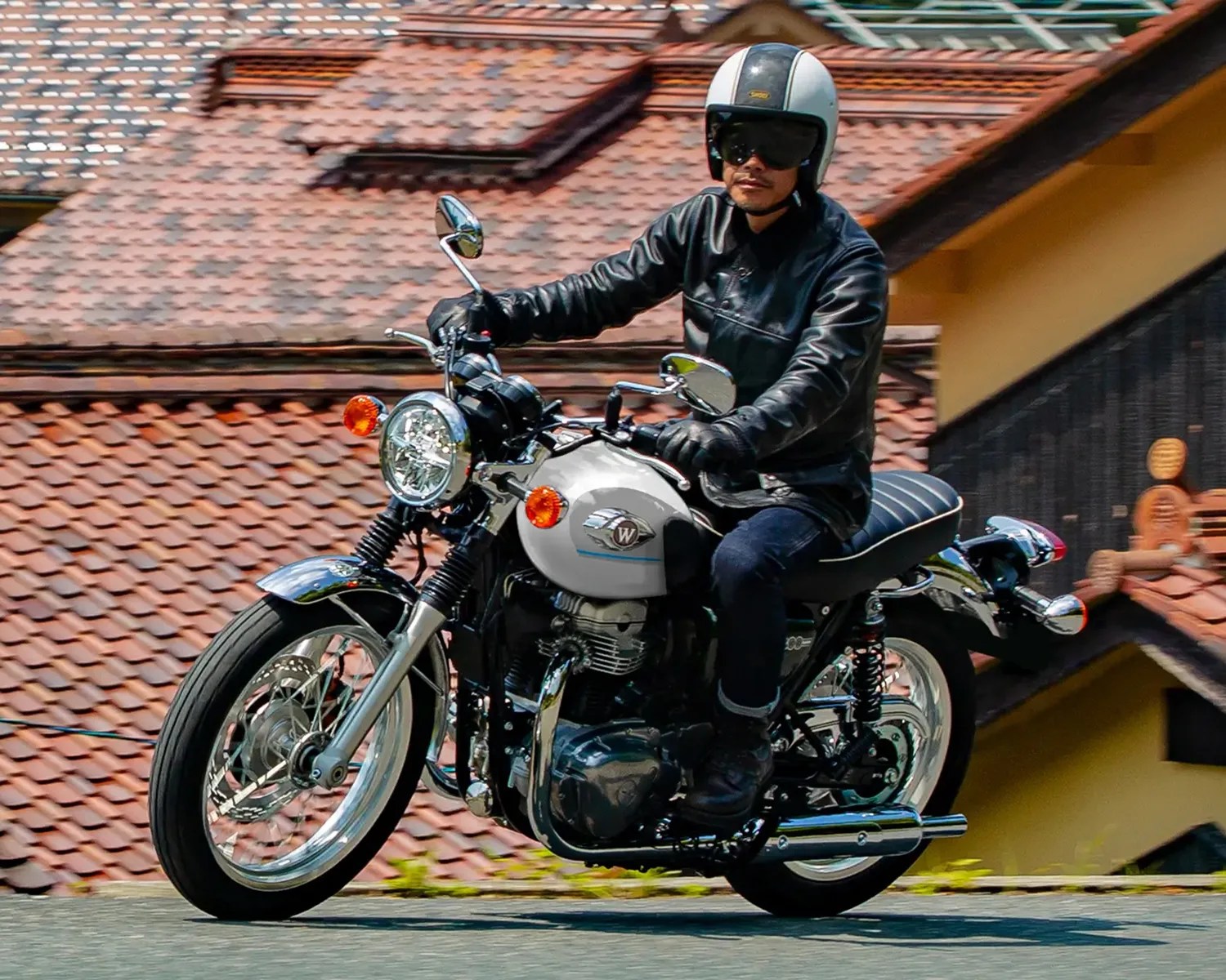 Person wearing a white helmet and black leather jacket riding a silver and black classic-style motorcycle on a road.