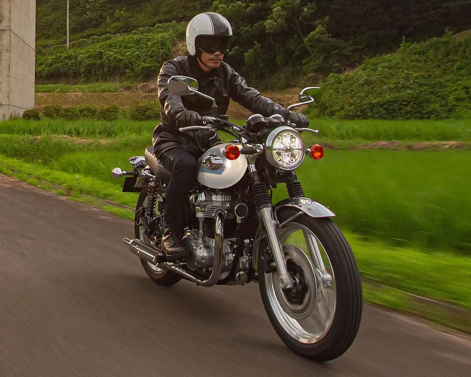 Person wearing a black leather jacket and white helmet riding a classic white and chrome motorcycle on a road.