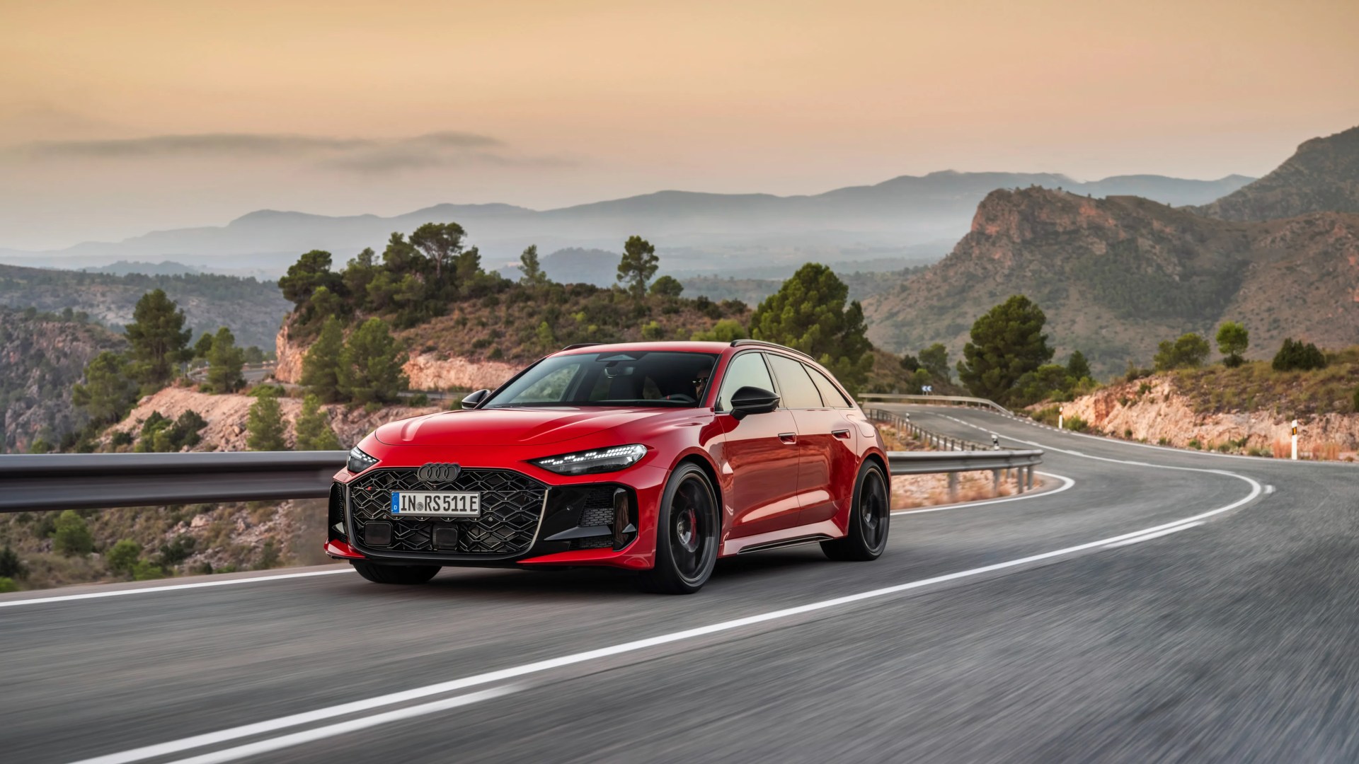 Red Audi RS 7 Avant with black grille and wheels driving on a winding mountain road at sunset.