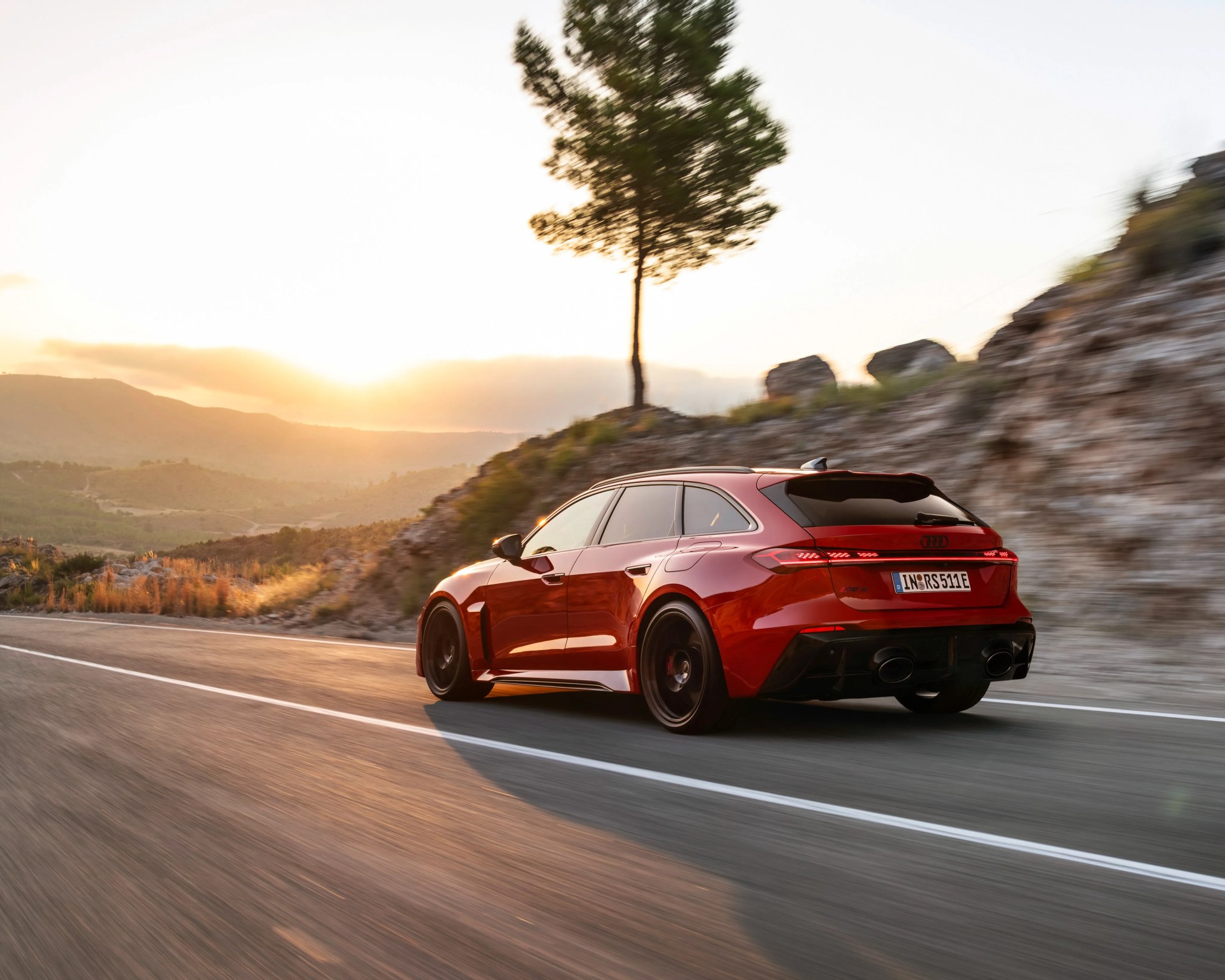 Red Audi RS Avant driving on a mountain road at sunset with rocky hills and a tree in the background.