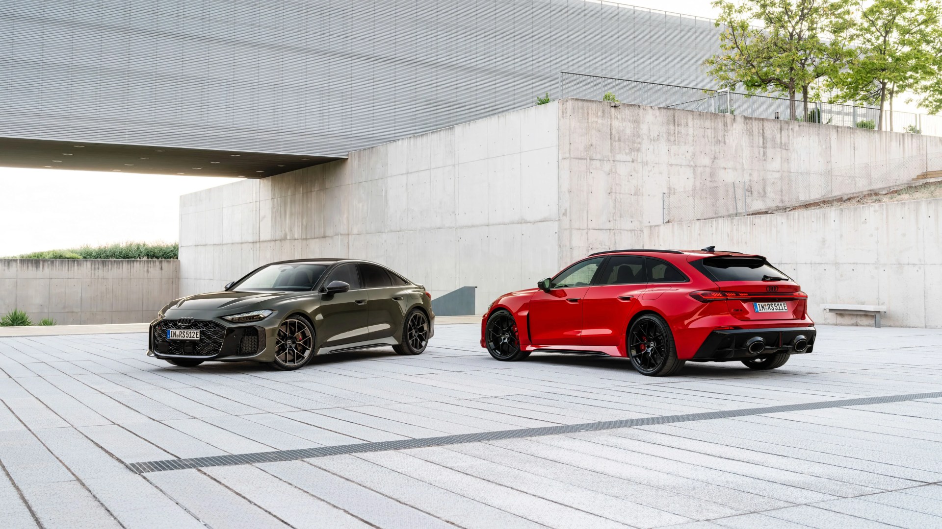 Two Audi RS models, one dark green sedan and one red wagon, parked on a light gray tiled surface near concrete walls.