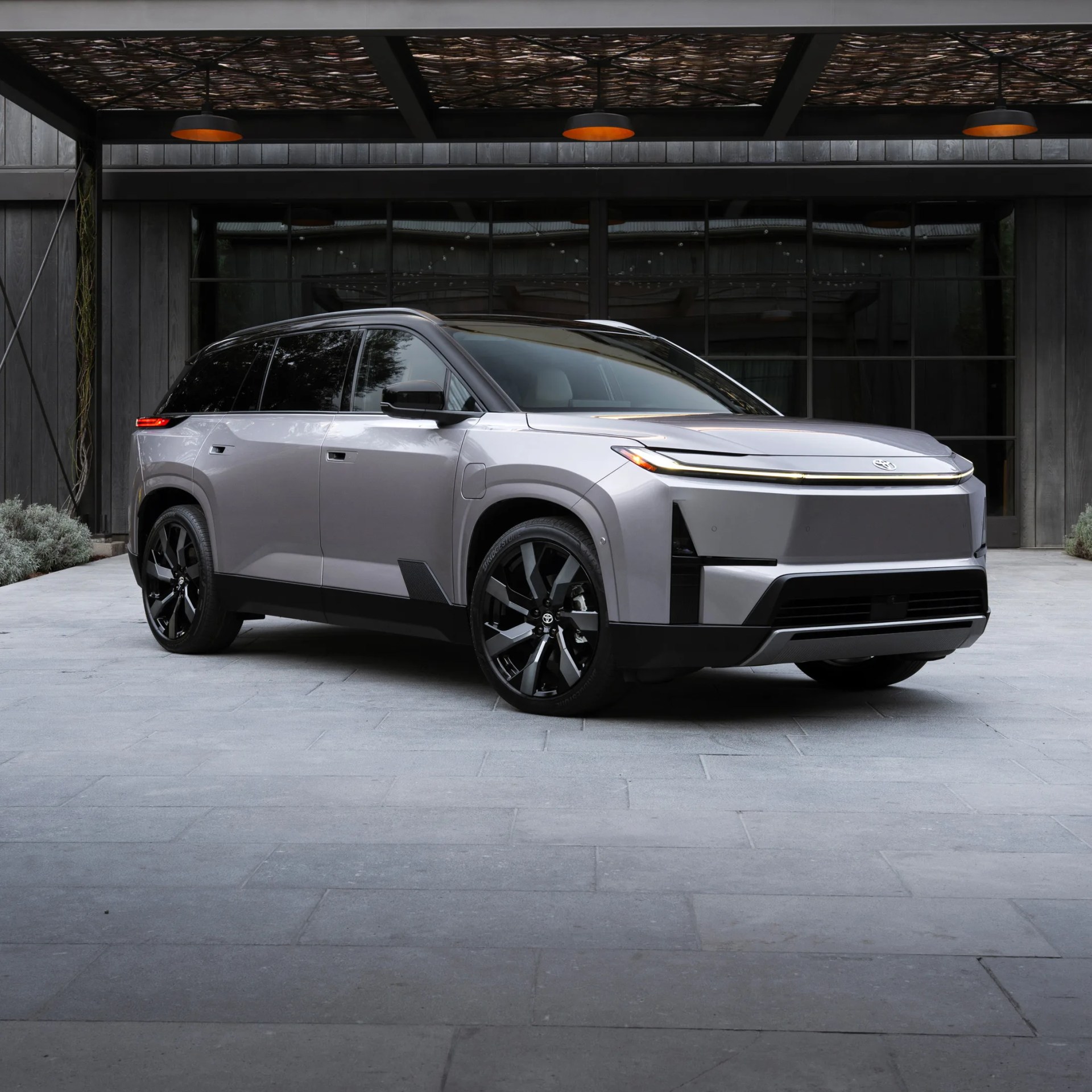 Silver electric SUV with black roof and wheels parked under a modern pergola with hanging lights.
