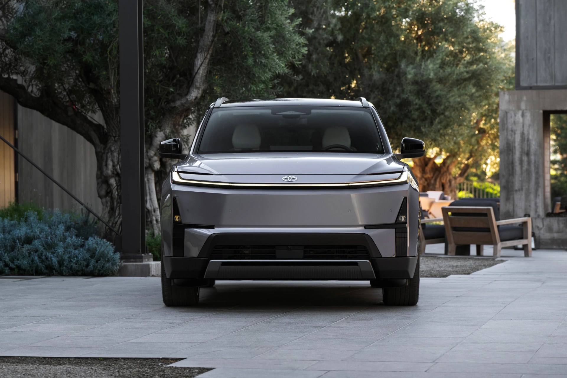 Front view of a silver Toyota electric SUV parked on a stone driveway with trees and outdoor furniture in the background.
