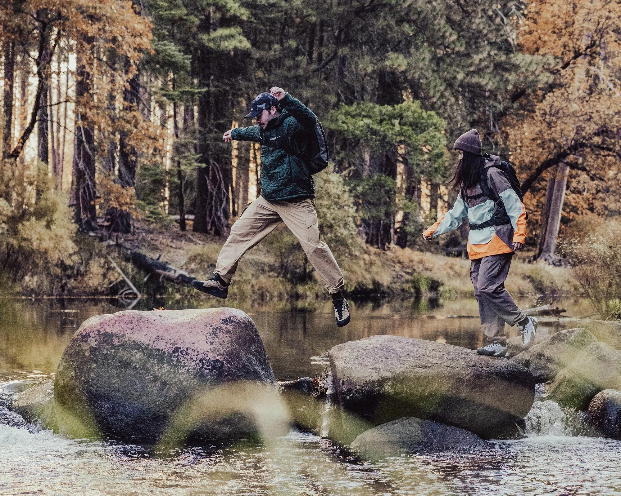 Two people wearing outdoor jackets and backpacks jumping across large rocks in a forest stream with autumn foliage.