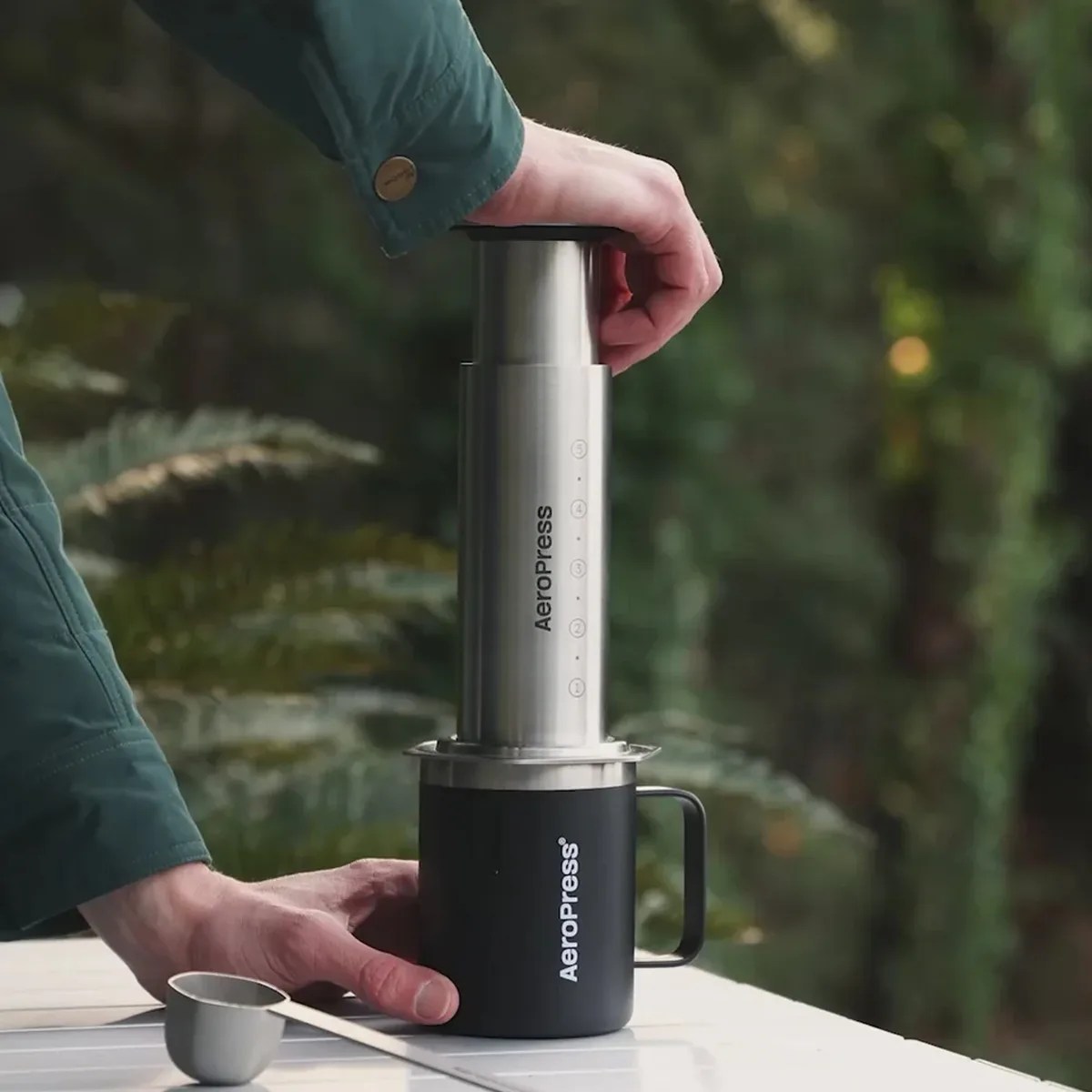 Person using a stainless steel AeroPress coffee maker on a black AeroPress mug outdoors.