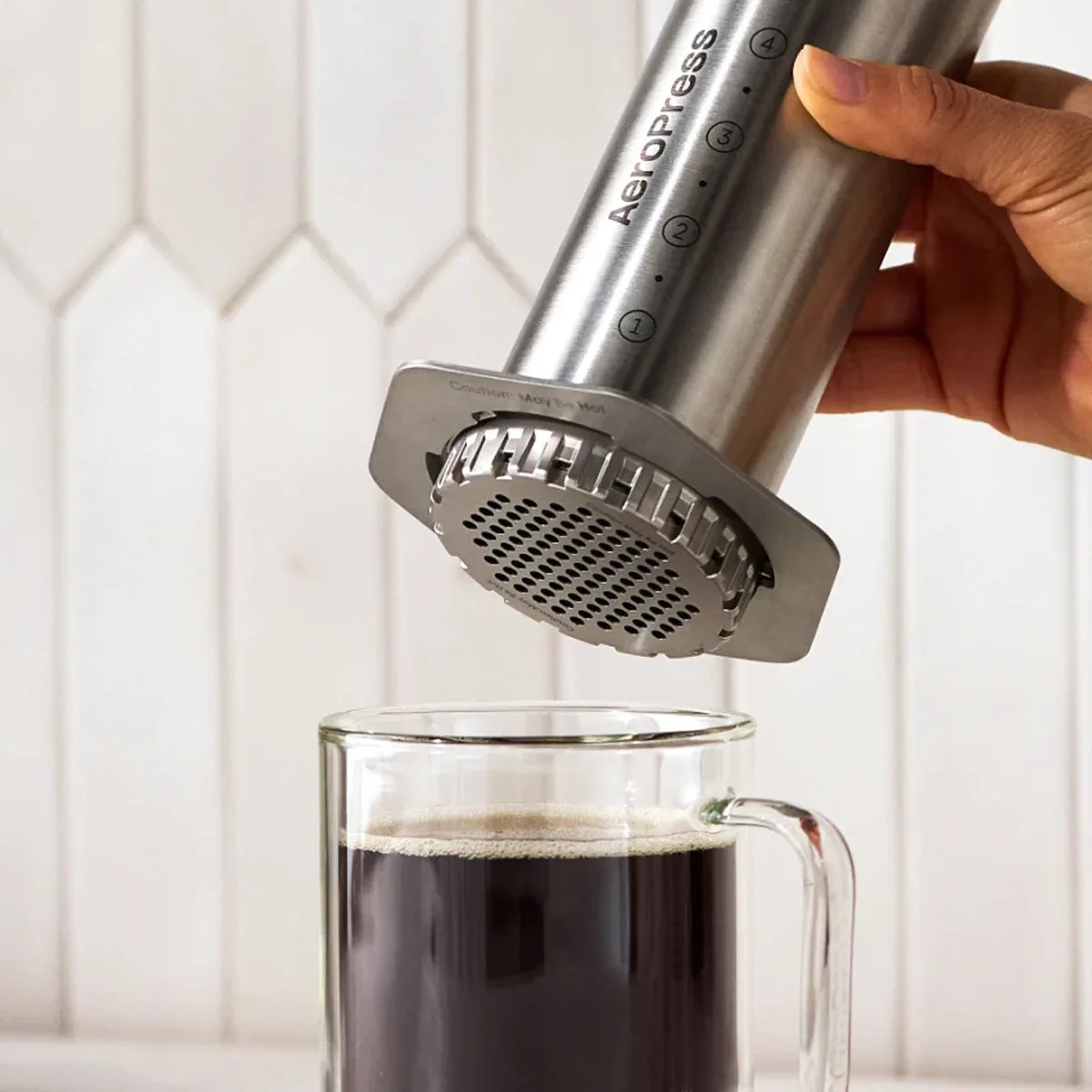 Hand holding a stainless steel AeroPress coffee maker above a glass mug filled with black coffee.