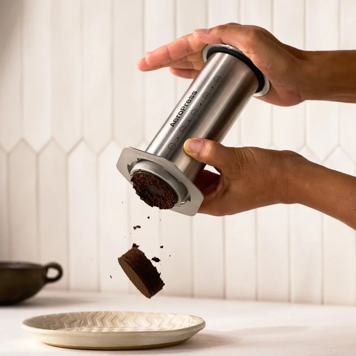 Hands pressing used coffee grounds out of a stainless steel AeroPress coffee maker over a beige plate.