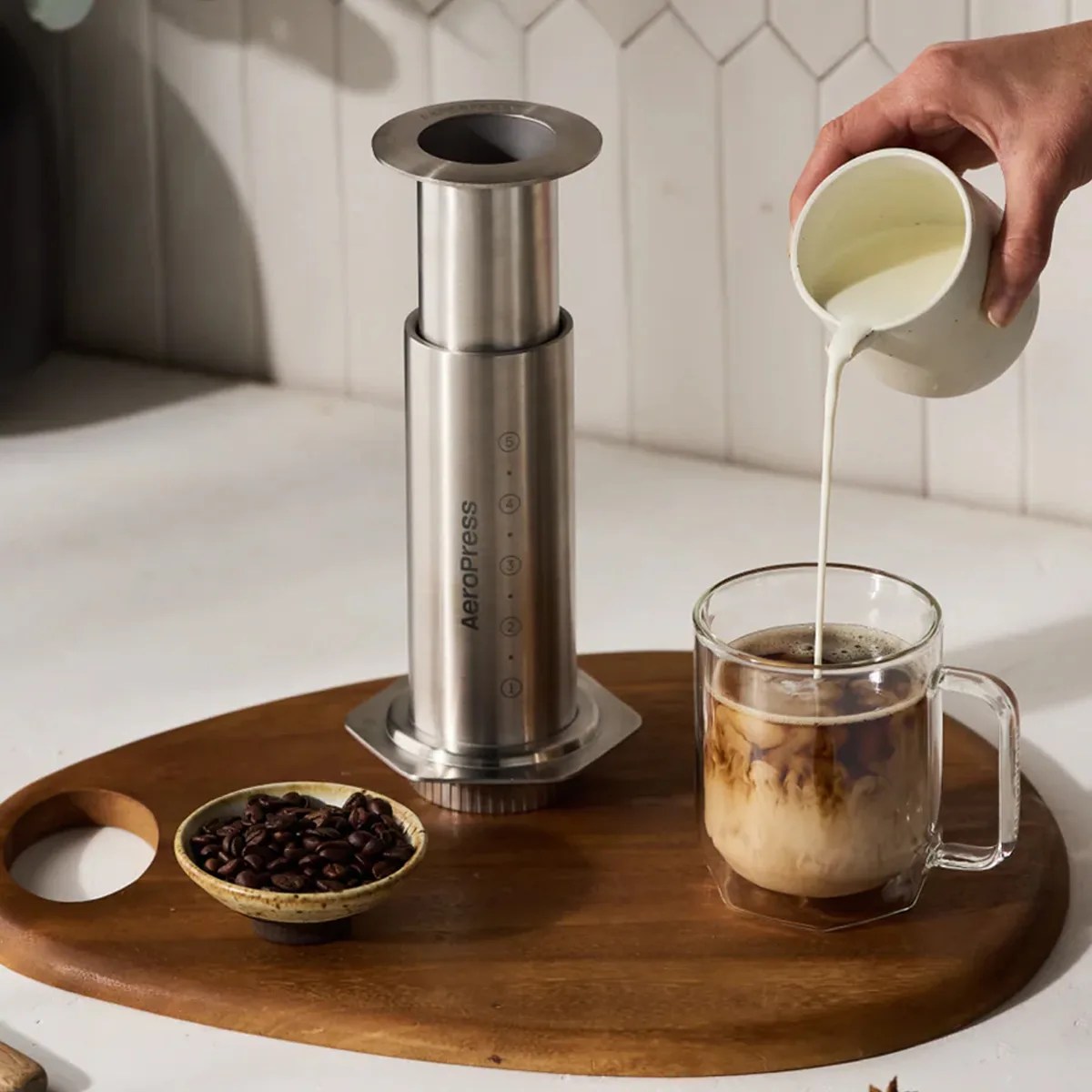 Stainless steel AeroPress coffee maker, glass mug with coffee and cream being poured, and bowl of coffee beans on wooden tray.