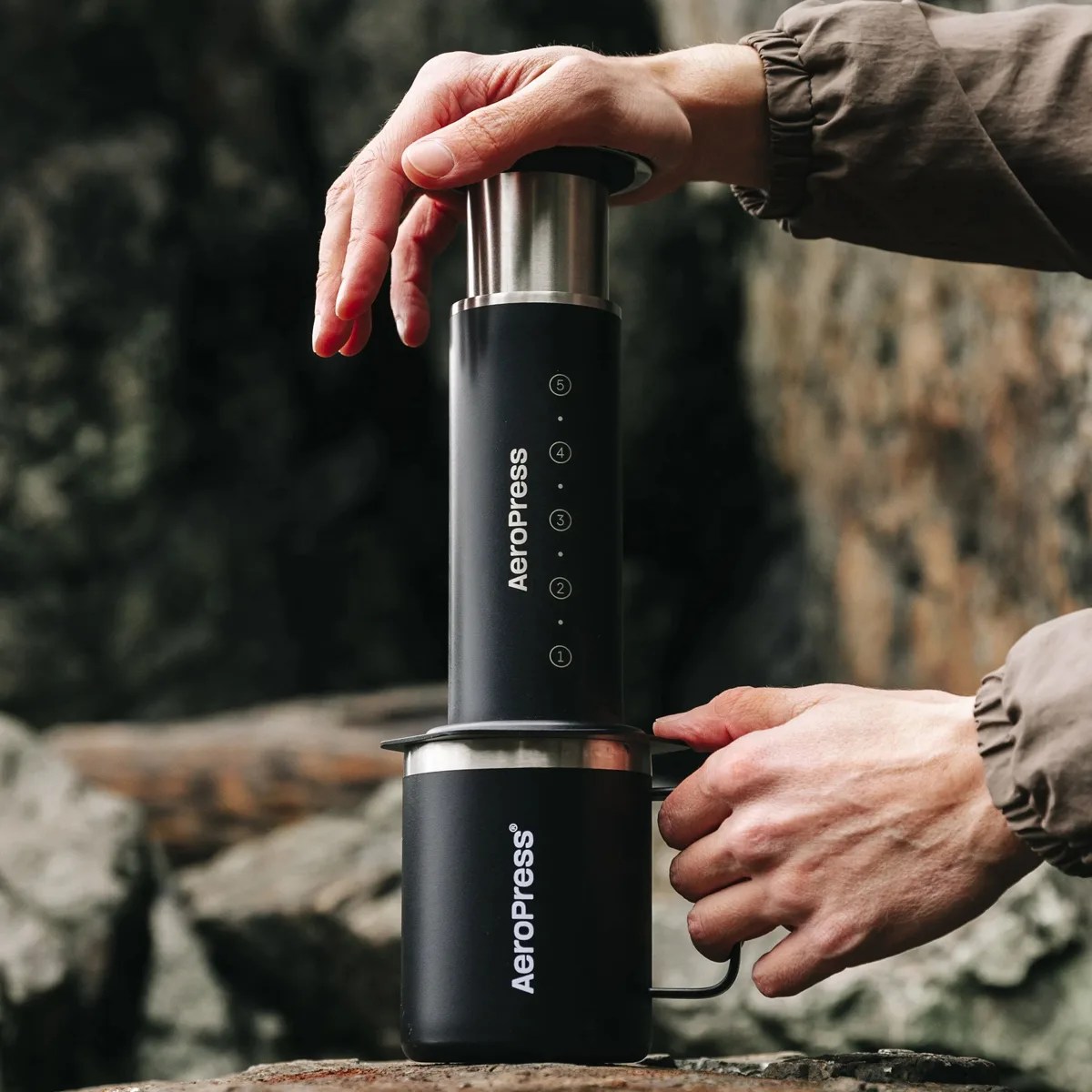 Black AeroPress coffee maker being used with a matching black AeroPress mug on a rocky surface.