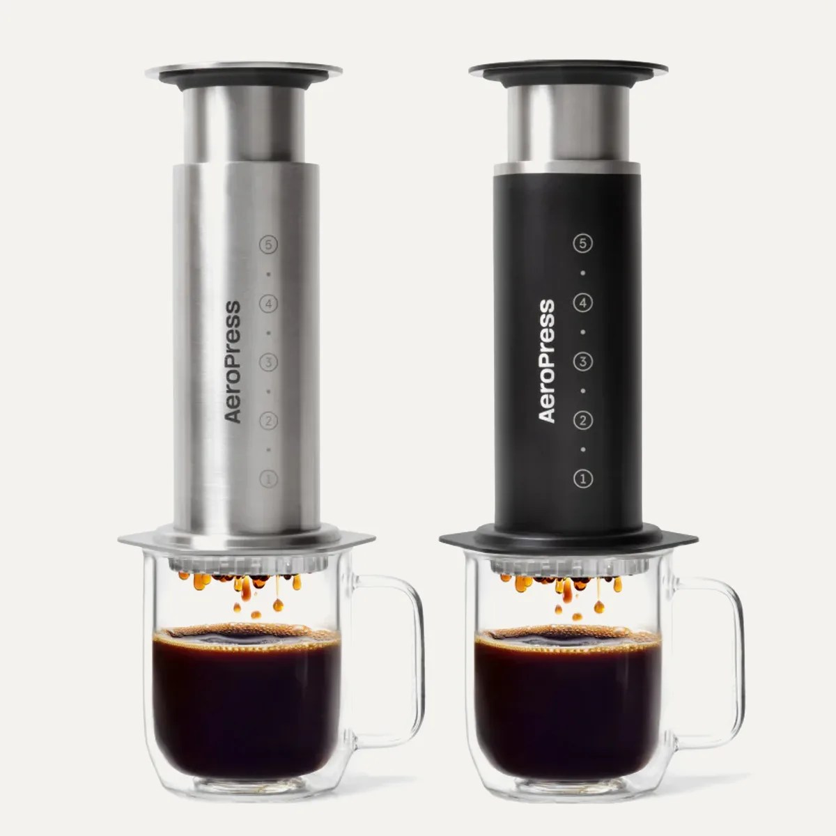 Two AeroPress coffee makers, one stainless steel and one black, brewing coffee into clear glass mugs.