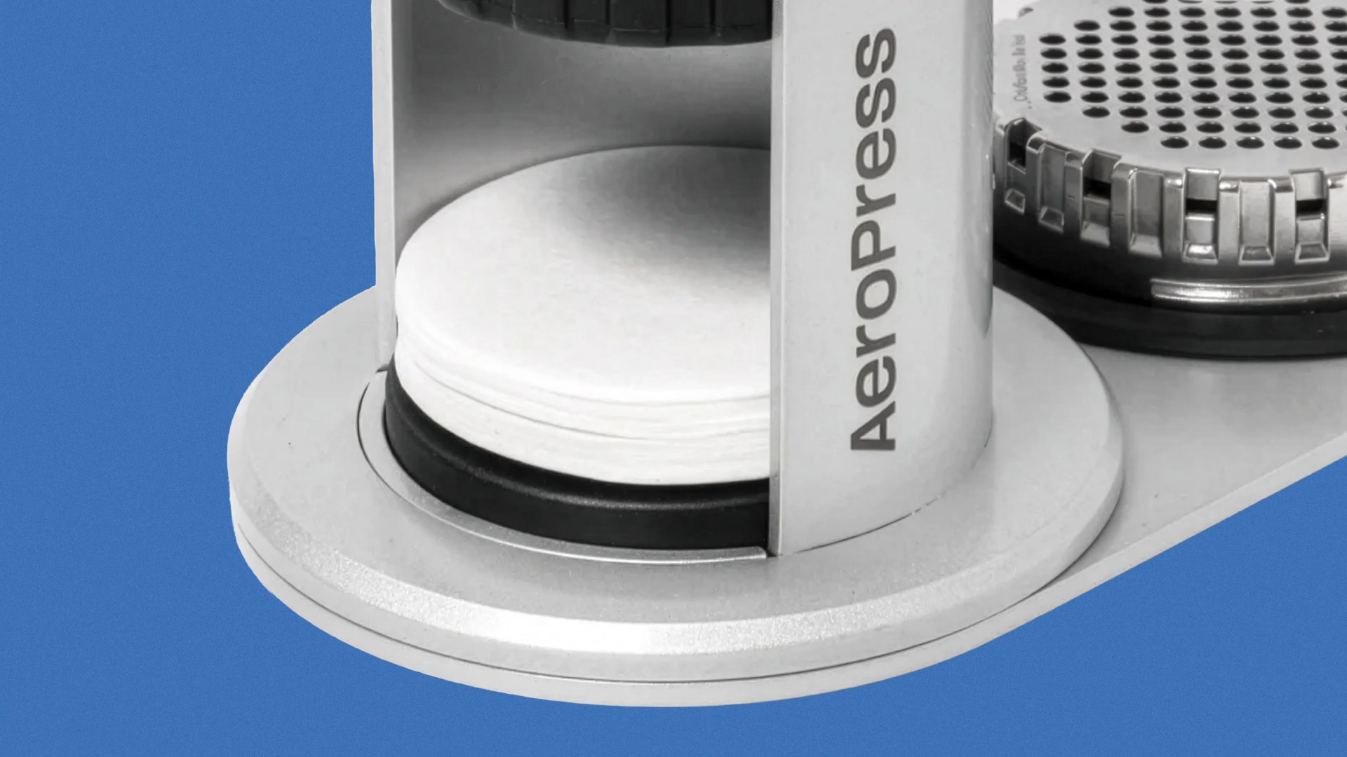 Close-up of a silver AeroPress coffee maker base holding white paper filters on a blue background.