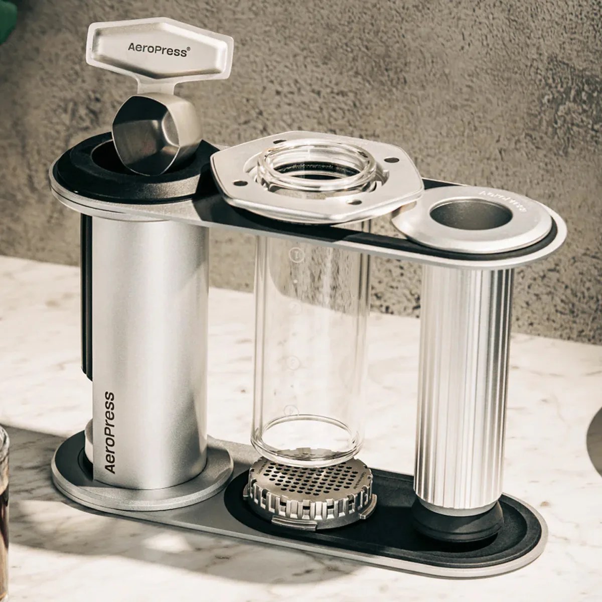 Silver and black AeroPress coffee maker with a clear cylindrical chamber and a scoop on a marble surface.
