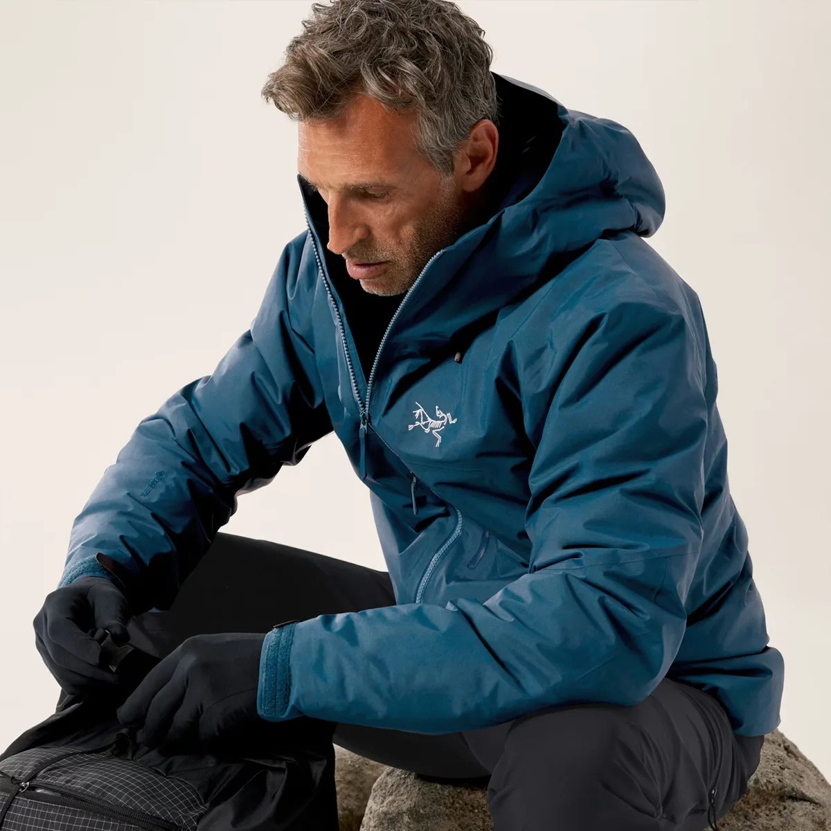 Man wearing a blue hooded jacket with a white logo, black gloves, and black pants while handling a black backpack.