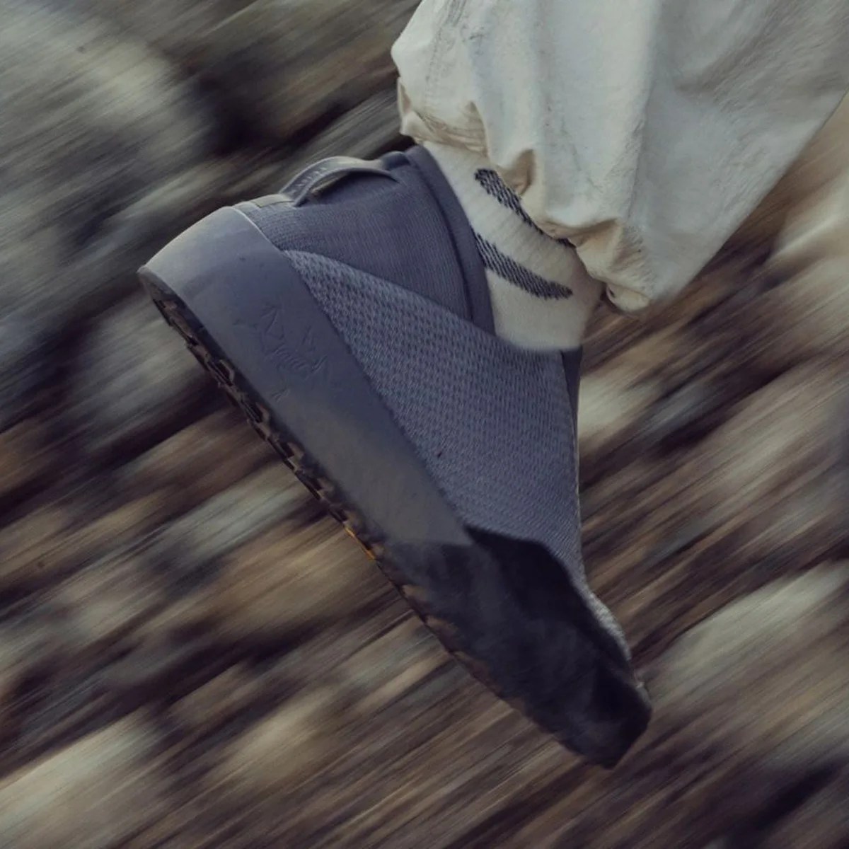 Gray slip-on sneaker with textured fabric and black toe cap worn with white pants and striped socks.