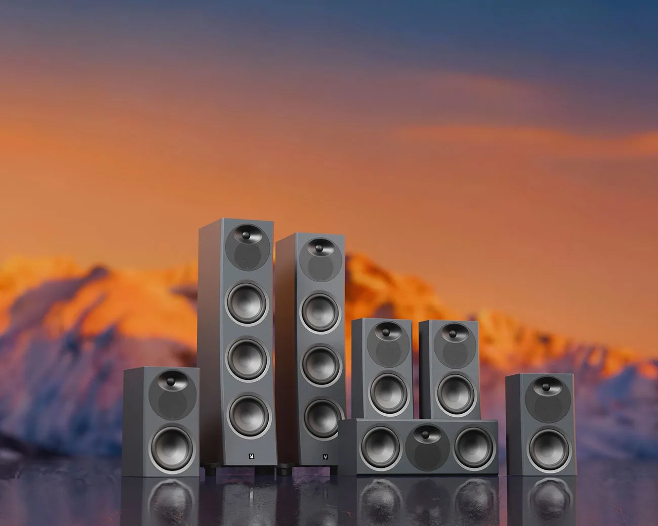 Set of six gray home theater speakers with multiple drivers, arranged on a reflective surface against a sunset mountain backdrop.