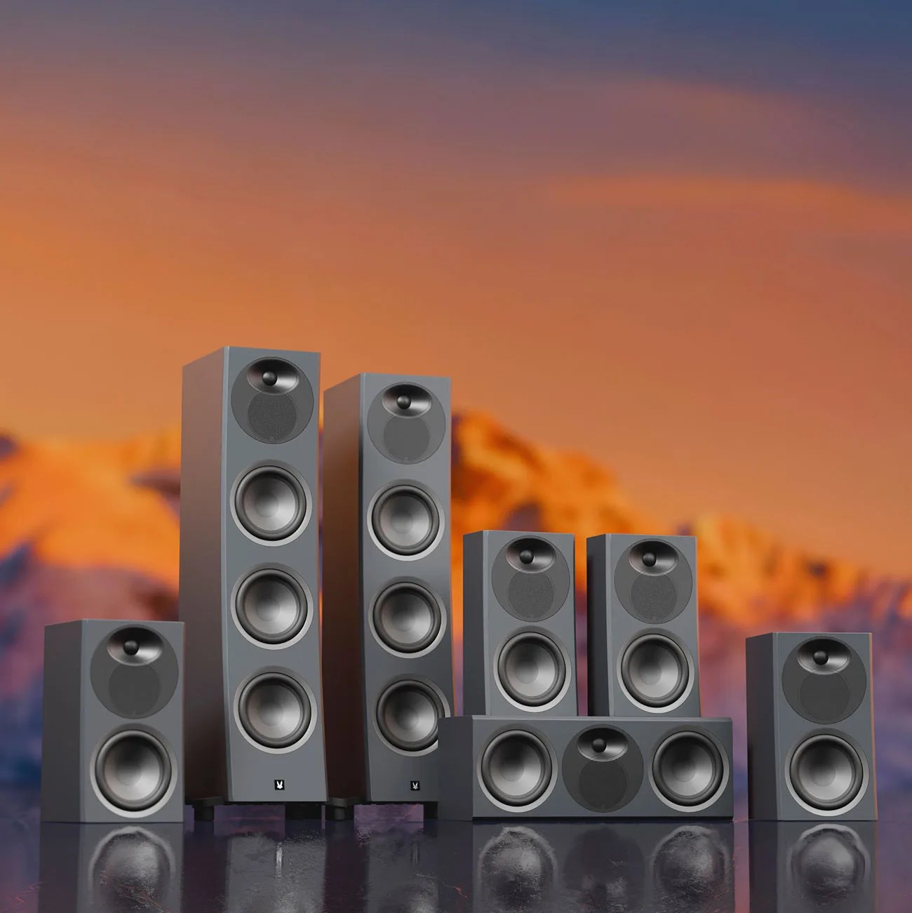 Set of six gray tower and bookshelf speakers with black speaker cones on a reflective surface against a sunset mountain background.