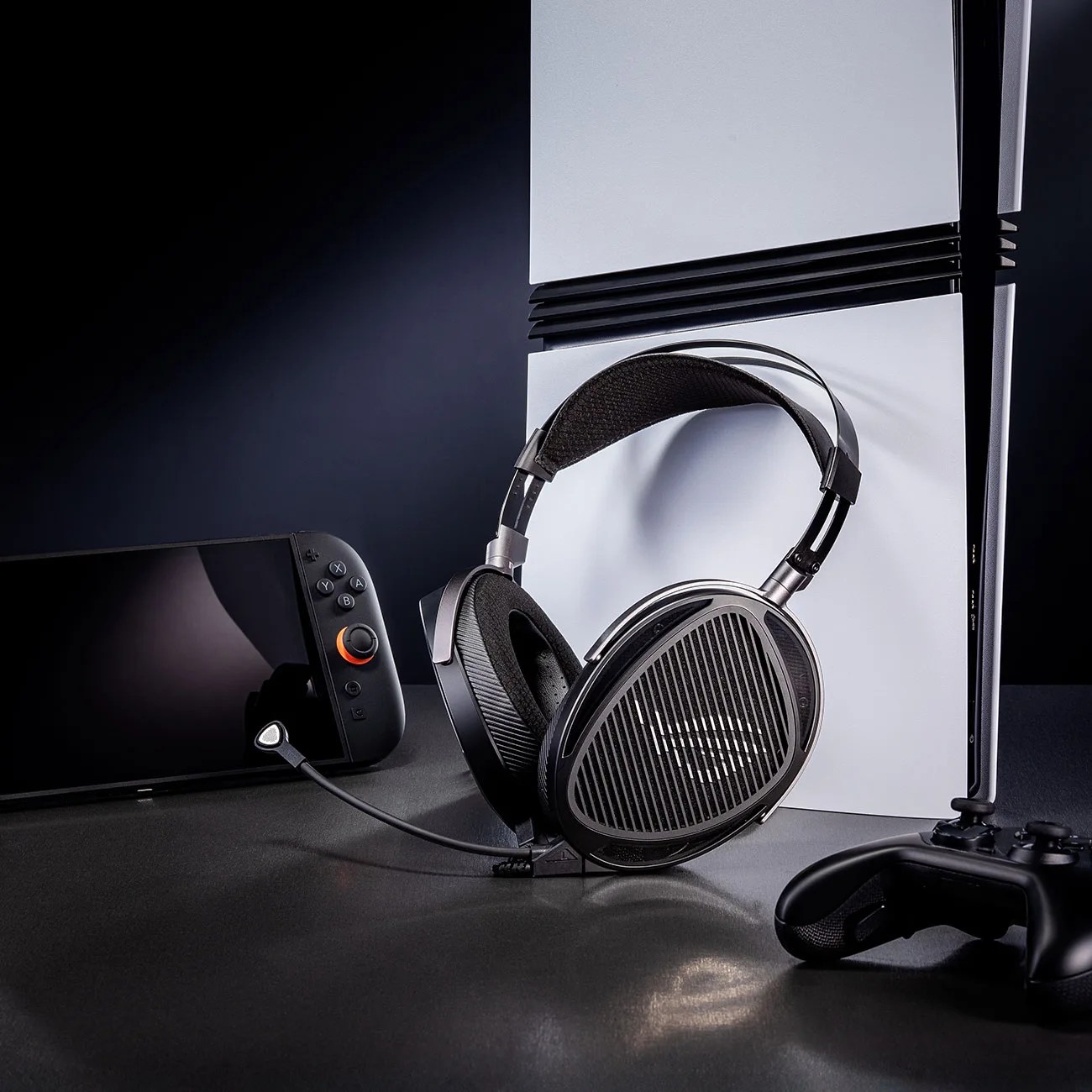 Black gaming headset with a microphone attached, placed in front of a white gaming console, next to a handheld console and a black controller.