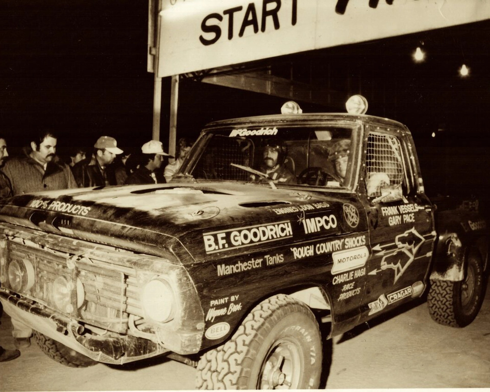 Vintage off-road race truck with sponsor decals including B.F. Goodrich and Motorola, under a "START" sign at night.