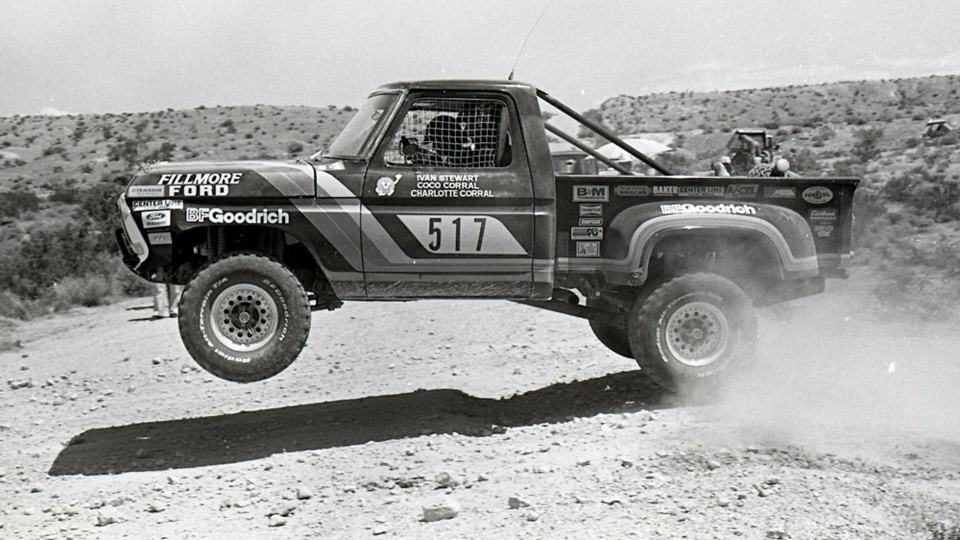 Off-road racing truck number 517 airborne over a dirt track with sponsor decals and a roll cage.