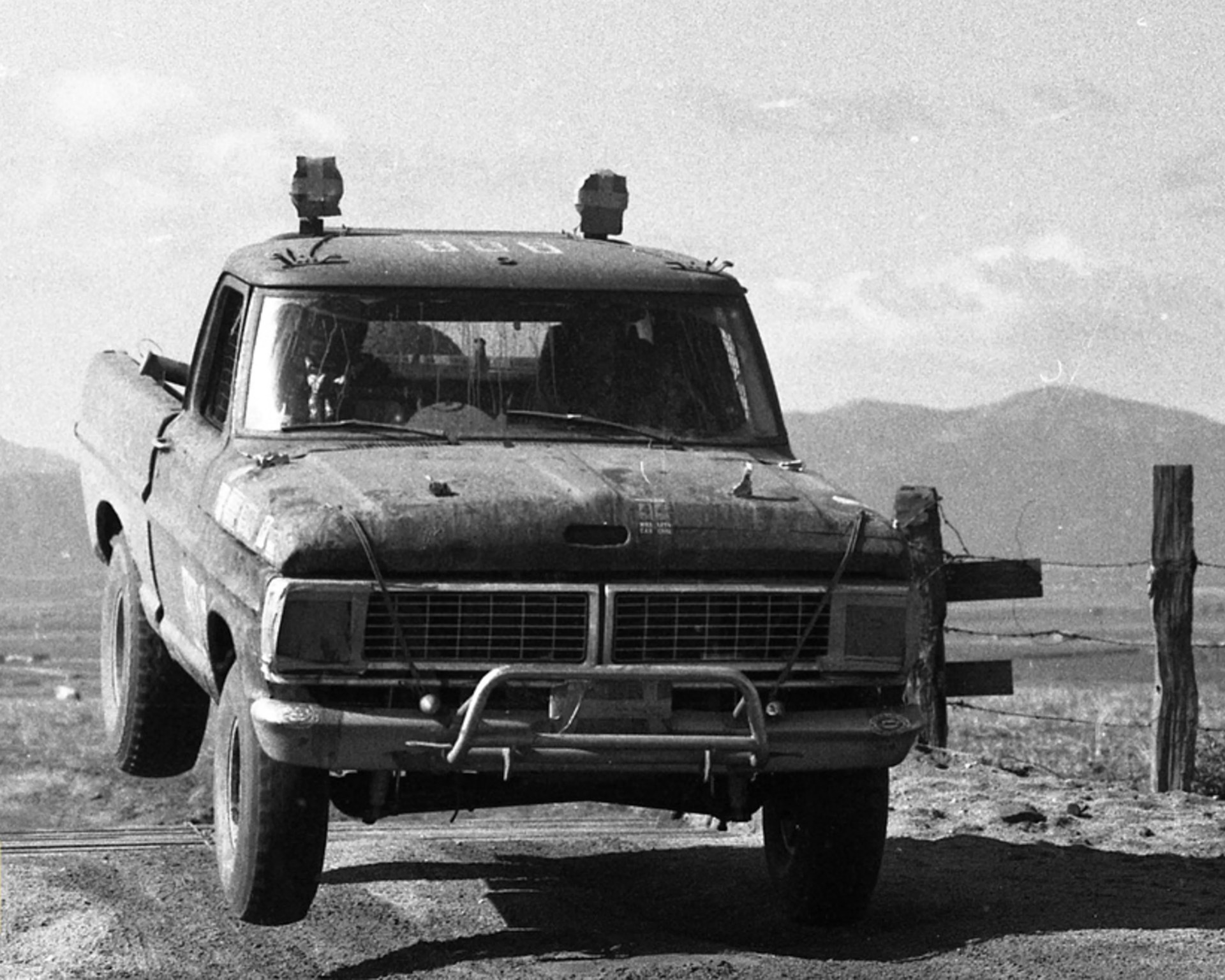 Black and white photo of an old, rugged pickup truck with front wheels off the ground on a dirt road near a barbed wire fence.