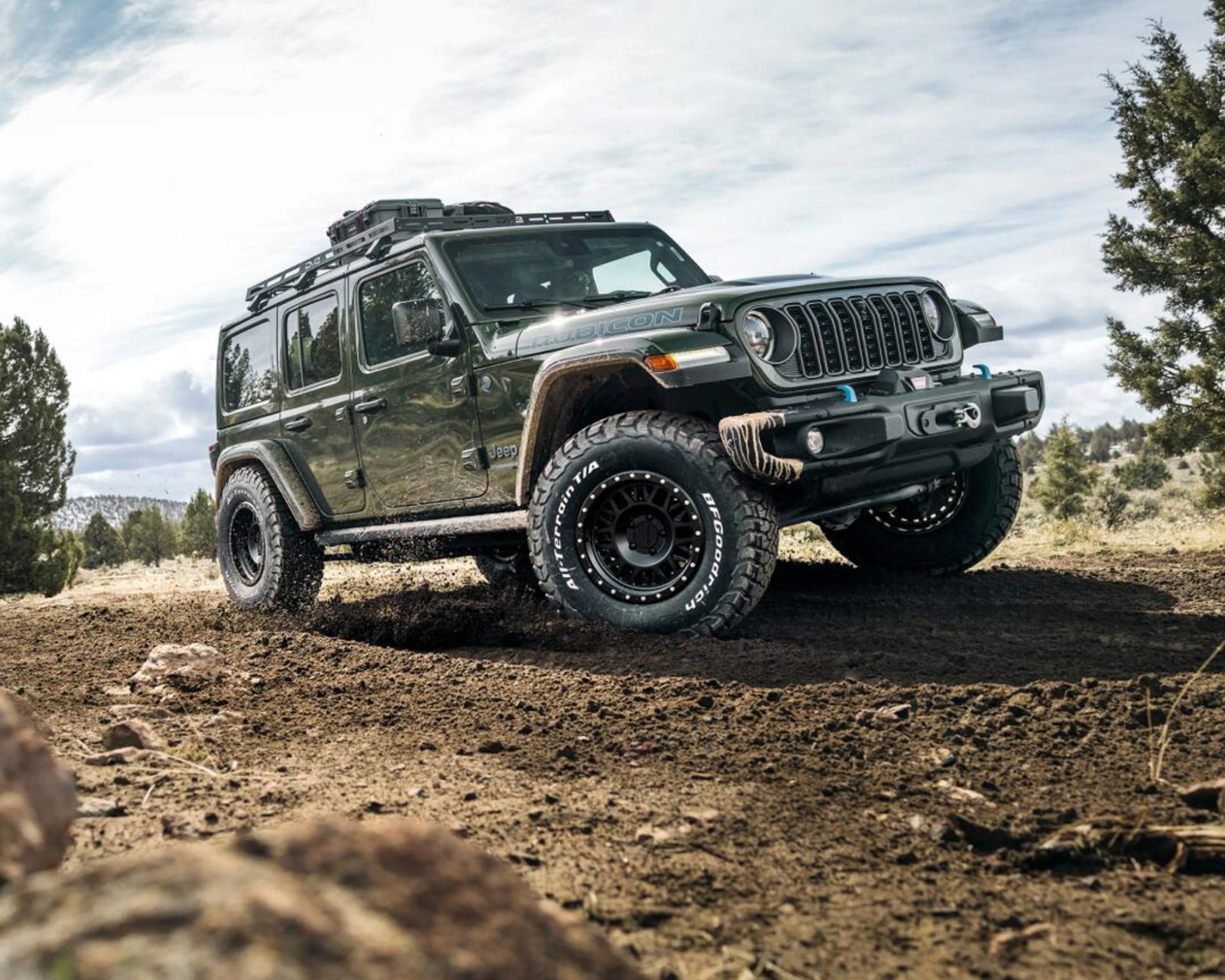 Dark green Jeep Rubicon with off-road tires driving on muddy terrain with trees in the background.