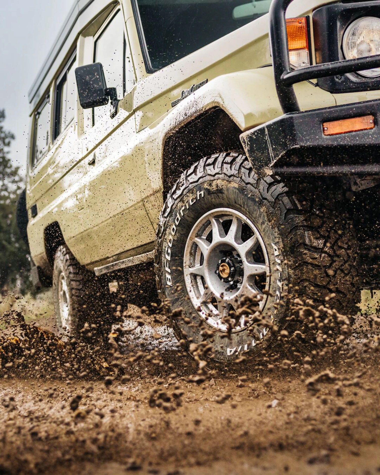 Beige Toyota Land Cruiser with BFGoodrich off-road tires splashing mud on a dirt trail.