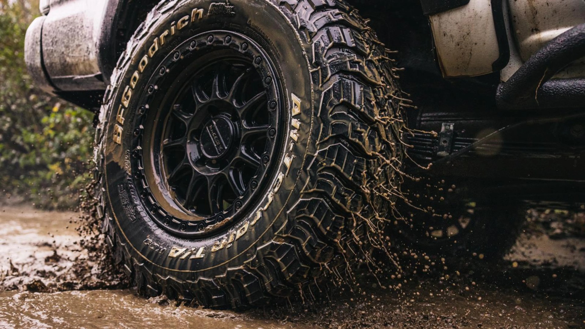 Black BFGoodrich All-Terrain T/A tire with aggressive tread splashing mud on a white off-road vehicle.