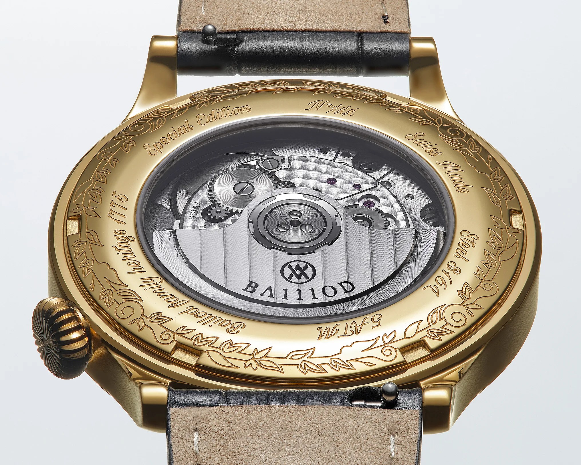 Gold watch case back with engraved floral design and text, showing mechanical movement through a transparent window.