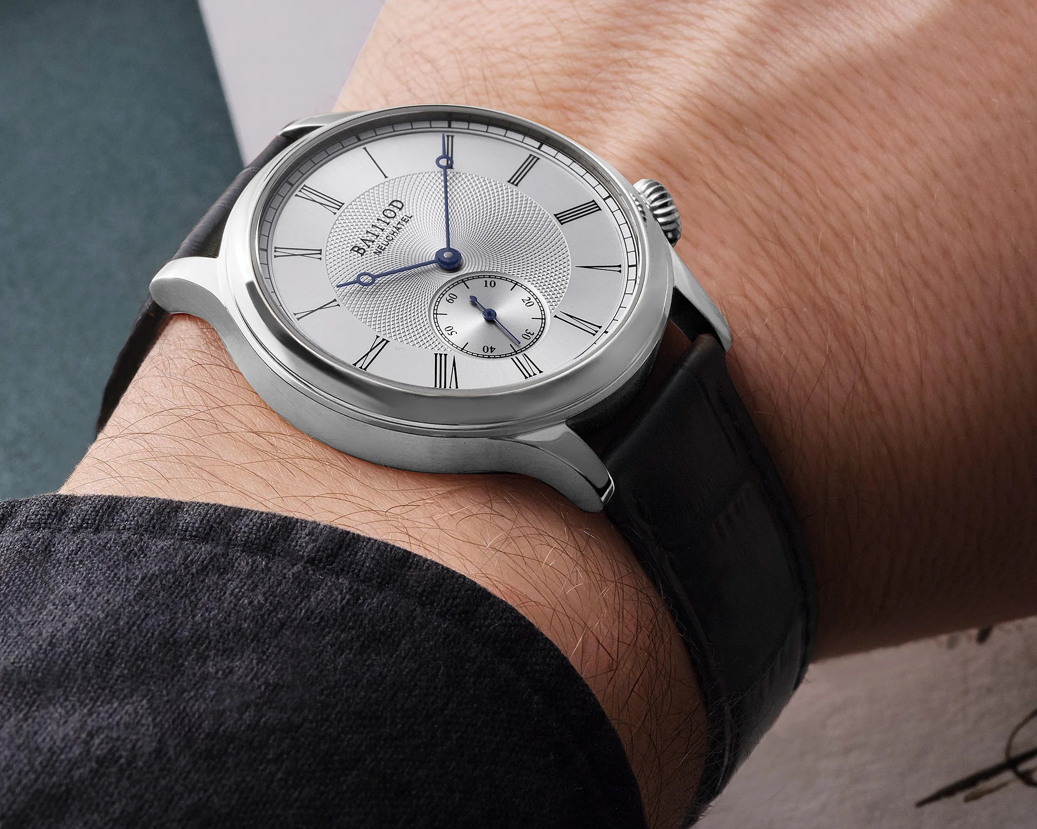 Silver Balmod wristwatch with Roman numerals and blue hands on a black leather strap worn on a wrist.