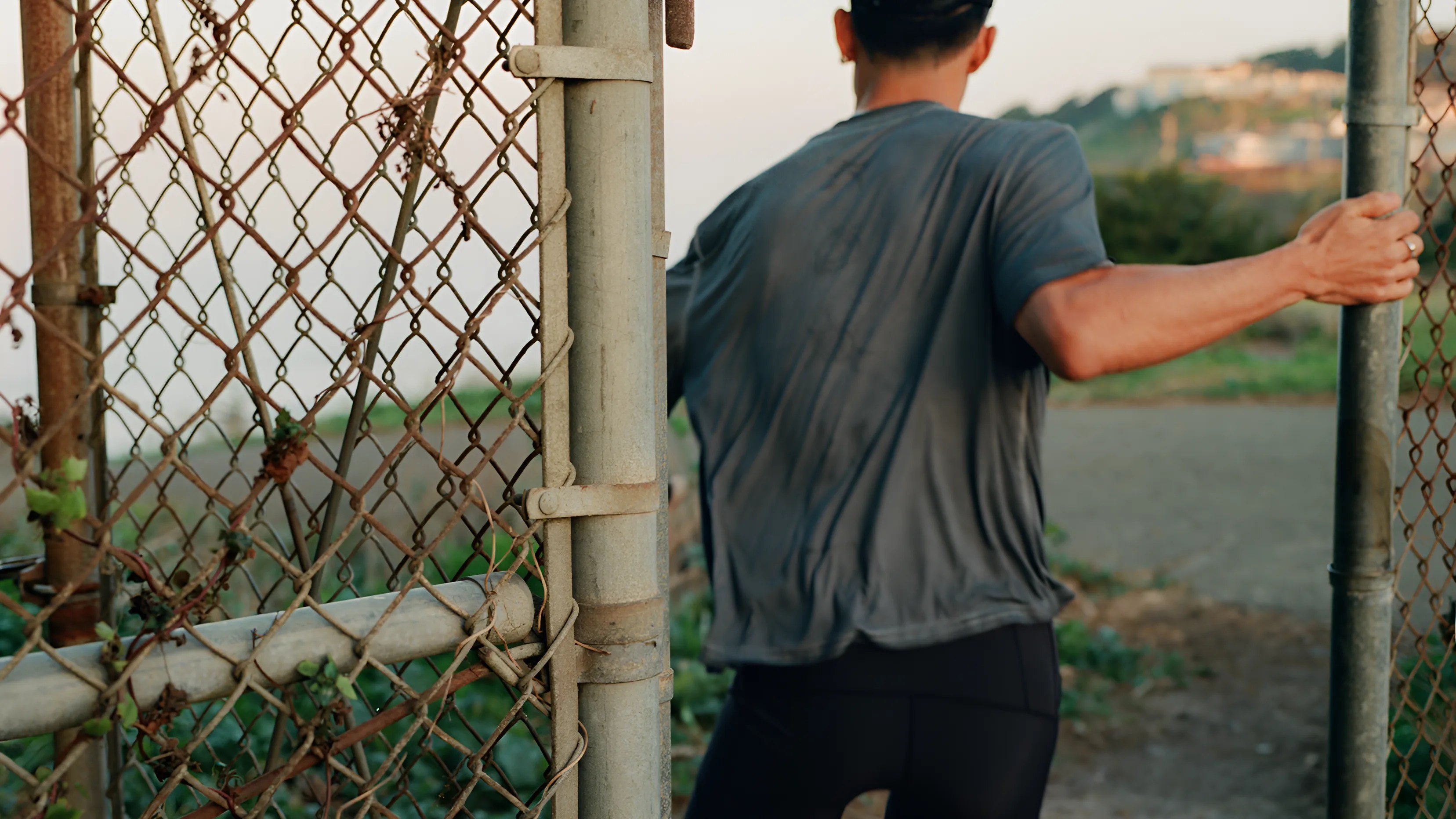 Person in a wet dark gray shirt and black pants opening a rusty chain-link gate outdoors.