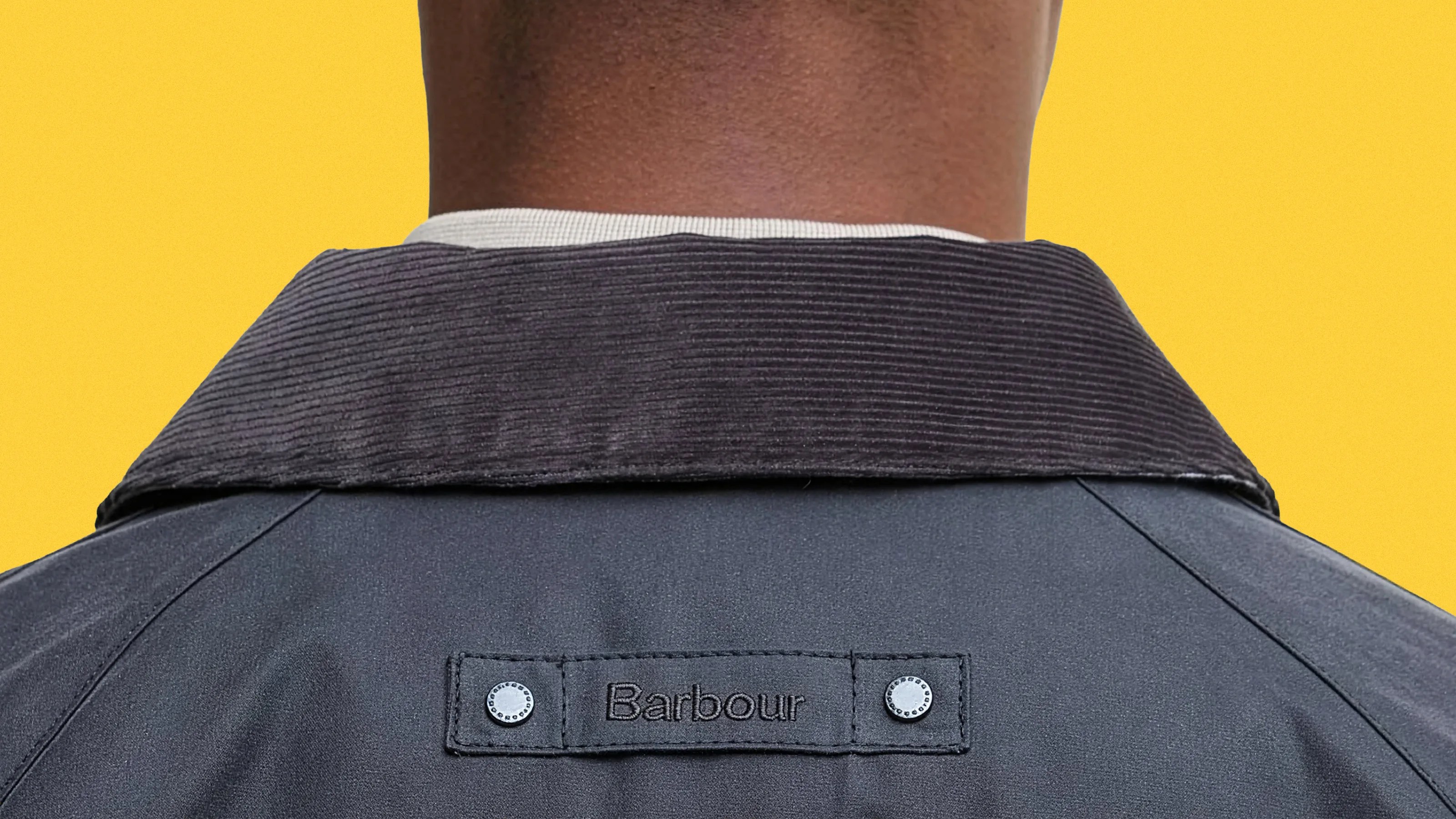 Back view of a person wearing a dark Barbour jacket with a corduroy collar against a yellow background.