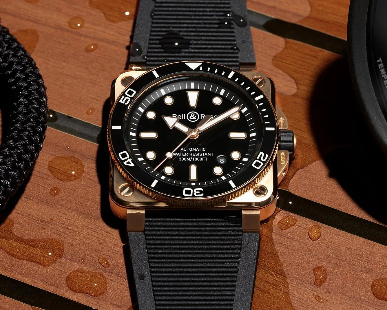 Square rose gold Bell & Ross dive watch with black dial, white markers, and black rubber strap on wet wooden surface.