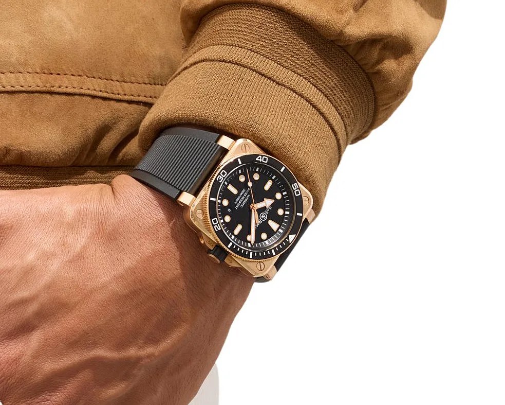 Square rose gold watch with black dial and bezel, worn on a wrist with a brown jacket sleeve.
